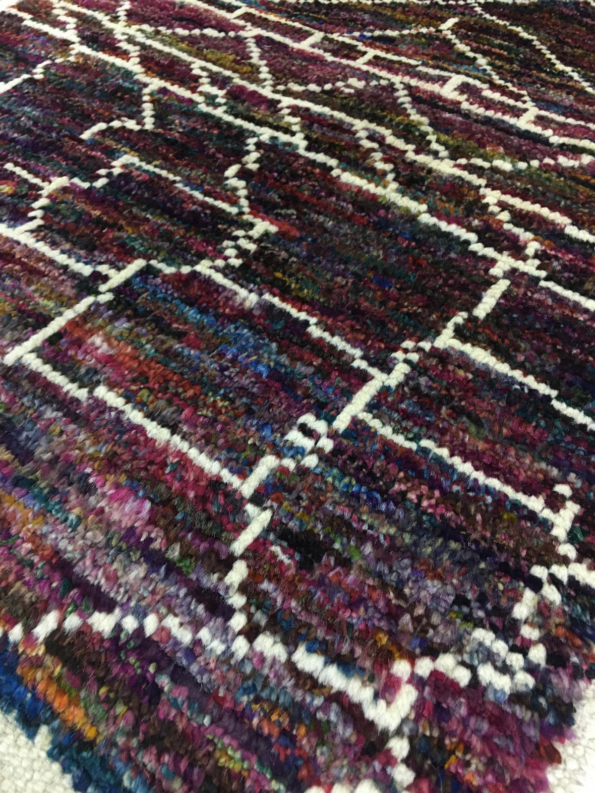 A close-up of a textured, multicolored area rug with a geometric diamond lattice pattern. The rug features a dark, heathered base of purple, blue, and red yarns outlined by thick white lines. Atlantica Organics curates antique and handmade rugs, Persian, Afghan, Tibetan, Pakistani, Turkish Oushak, Kilim, Khotan, tribal and contemporary textiles for luxury interiors, homes and real estate. Our collections connect timeless craftsmanship with interior design, furniture, fine art, architecture and modern living