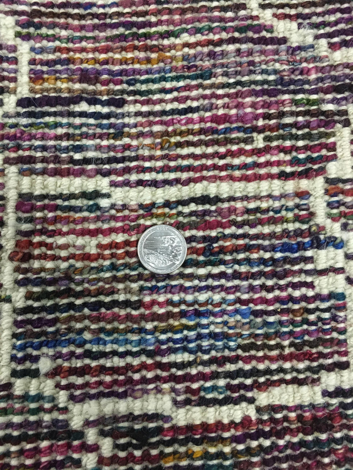 Close-up of a multicolored woven rug with a silver coin placed in the center for scale. Atlantica Organics curates antique and handmade rugs, Persian, Afghan, Tibetan, Pakistani, Turkish Oushak, Kilim, Khotan, tribal and contemporary textiles for luxury interiors, homes and real estate. Our collections connect timeless craftsmanship with interior design, furniture, fine art, architecture and modern living. Explore curated rugs, collectible textiles, natural materials, cultural heritage, sustainable design a
