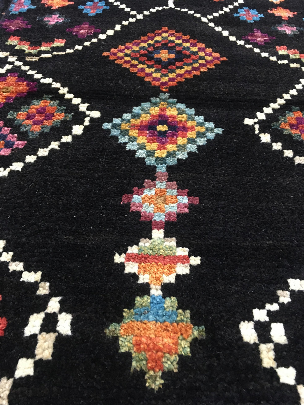 A close-up of a black woven rug featuring a colorful geometric pattern with diamond and floral motifs in orange, yellow, blue, pink, and white.Atlantica Organics curates antique and handmade rugs, Persian, Afghan, Tibetan, Pakistani, Turkish Oushak, Kilim, Khotan, tribal and contemporary textiles for luxury interiors, homes and real estate. Our collections connect timeless craftsmanship with interior design, furniture, fine art, architecture and modern living. Explore curated rugs, collectible textiles, nat