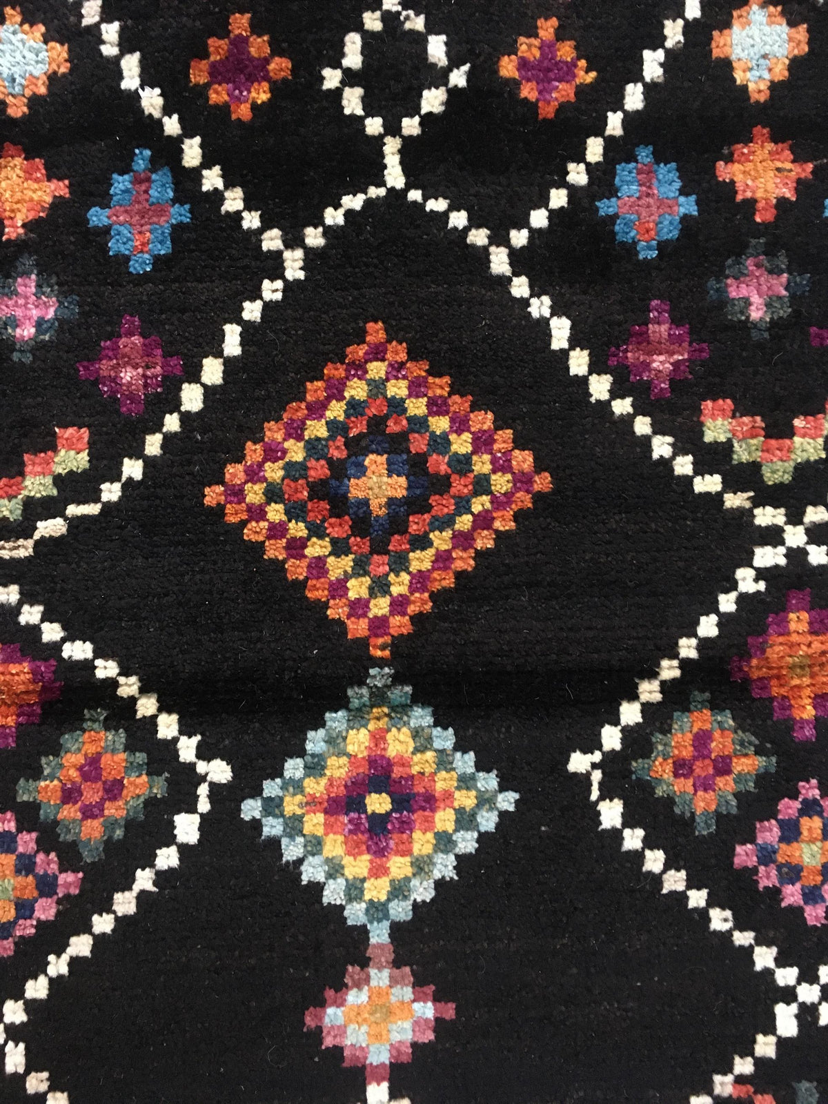 A close-up of a black textile or rug featuring a geometric diamond pattern in cream, orange, red, blue, and purple. Atlantica Organics curates antique and handmade rugs, Persian, Afghan, Tibetan, Pakistani, Turkish Oushak, Kilim, Khotan, tribal and contemporary textiles for luxury interiors, homes and real estate. Our collections connect timeless craftsmanship with interior design, furniture, fine art, architecture and modern living. Explore curated rugs, collectible textiles, natural materials, cultural he