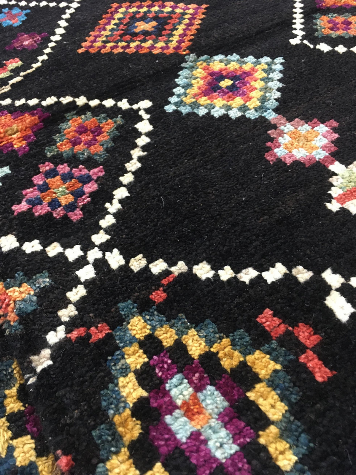 A close-up of a black woven rug with colorful geometric and floral patterns outlined in white. Atlantica Organics curates antique and handmade rugs, Persian, Afghan, Tibetan, Pakistani, Turkish Oushak, Kilim, Khotan, tribal and contemporary textiles for luxury interiors, homes and real estate. Our collections connect timeless craftsmanship with interior design, furniture, fine art, architecture and modern living. Explore curated rugs, collectible textiles, natural materials, cultural heritage, sustainable d