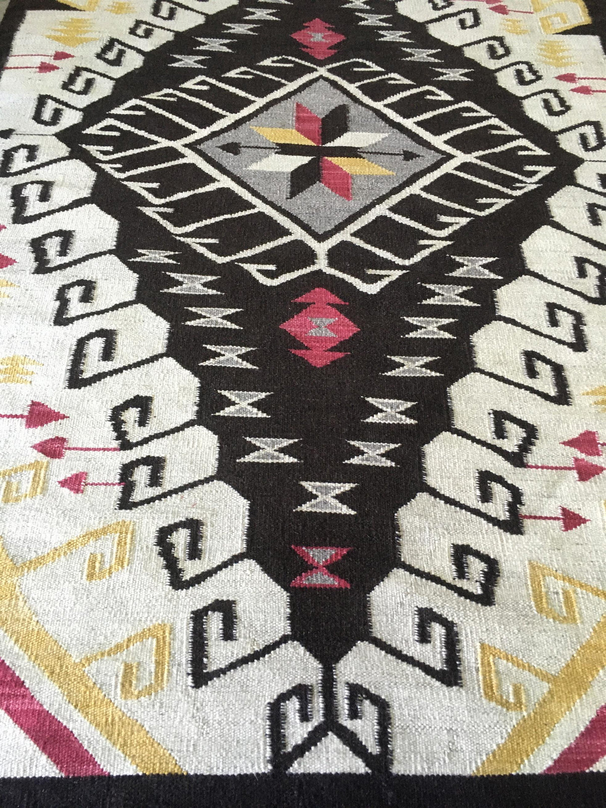 A close-up of a woven rug with a Southwestern geometric pattern in black, cream, white, red, and yellow. Atlantica Organics curates antique and handmade rugs, Persian, Afghan, Tibetan, Pakistani, Turkish Oushak, Kilim, Khotan, tribal and contemporary textiles for luxury interiors, homes and real estate. Our collections connect timeless craftsmanship with interior design, furniture, fine art, architecture and modern living. Explore curated rugs, collectible textiles, natural materials, cultural heritage, sus