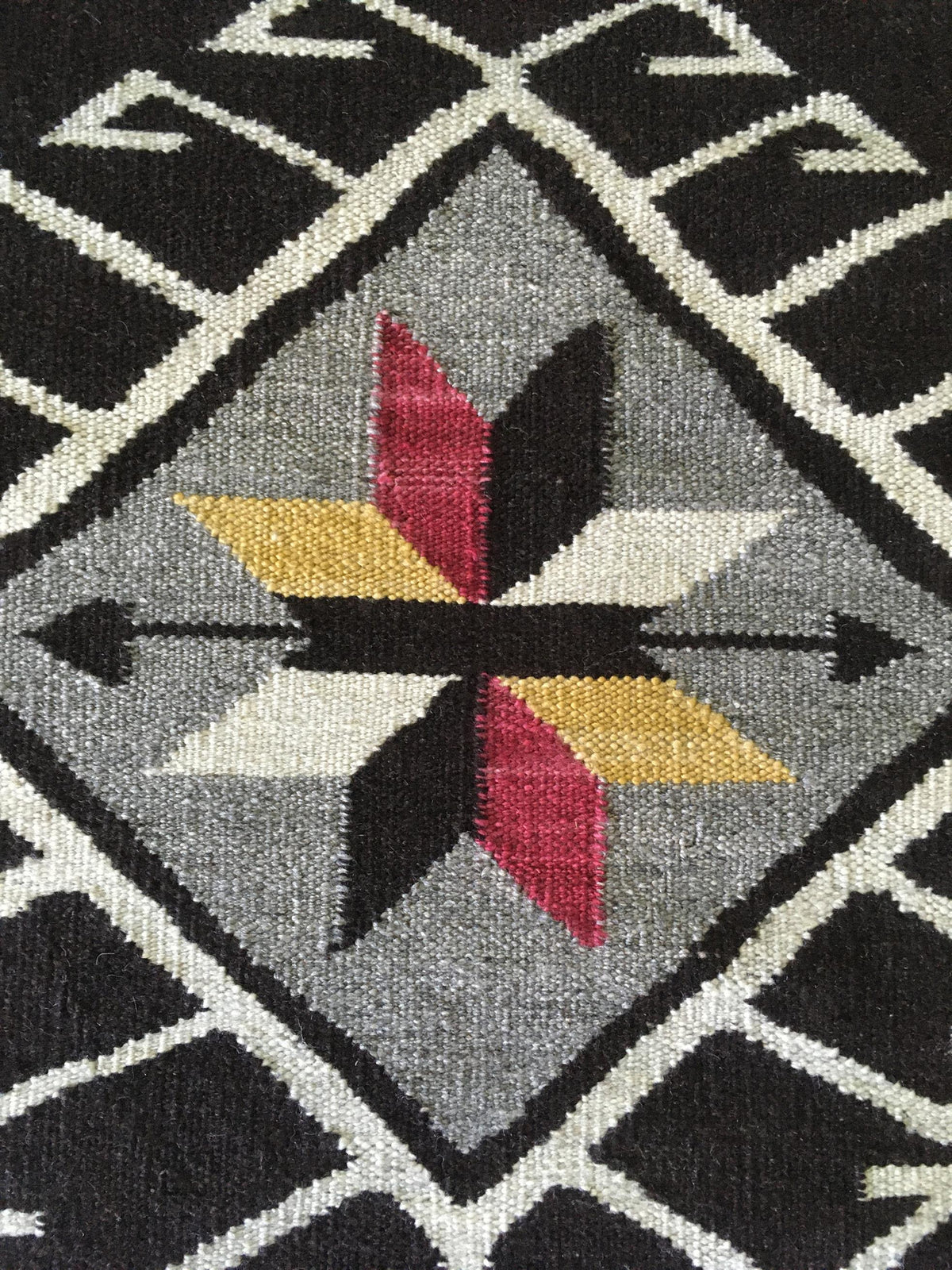 Close-up of a woven textile with a geometric Southwestern-style pattern, featuring a central diamond motif in gray, black, white, yellow, and red on a black background. Atlantica Organics curates antique and handmade rugs, Persian, Afghan, Tibetan, Pakistani, Turkish Oushak, Kilim, Khotan, tribal and contemporary textiles for luxury interiors, homes and real estate. Our collections connect timeless craftsmanship with interior design, furniture, fine art, architecture and modern living. Explore curated rugs,