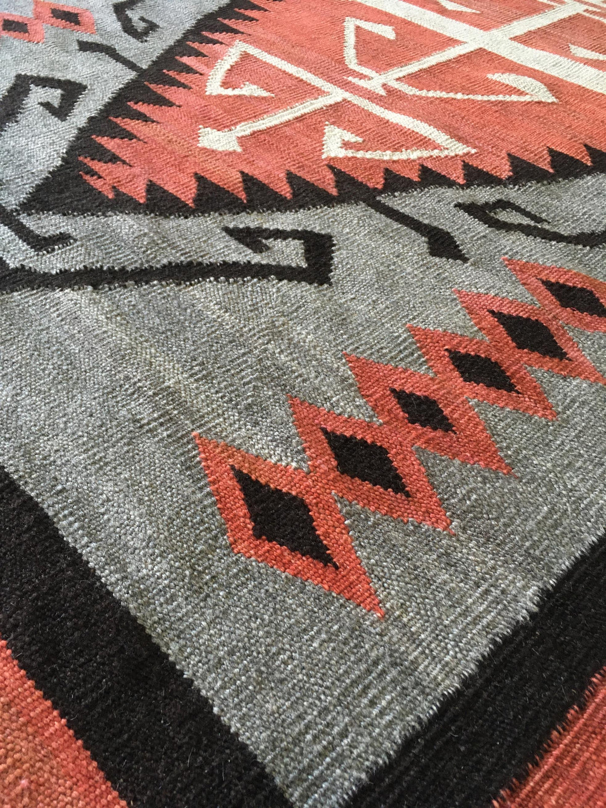 Close-up of a woven rug with a gray field, black geometric borders, and rust-red diamond and zigzag motifs. Atlantica Organics curates antique and handmade rugs, Persian, Afghan, Tibetan, Pakistani, Turkish Oushak, Kilim, Khotan, tribal and contemporary textiles for luxury interiors, homes and real estate. Our collections connect timeless craftsmanship with interior design, furniture, fine art, architecture and modern living. Explore curated rugs, collectible textiles, natural materials, cultural heritage, 
