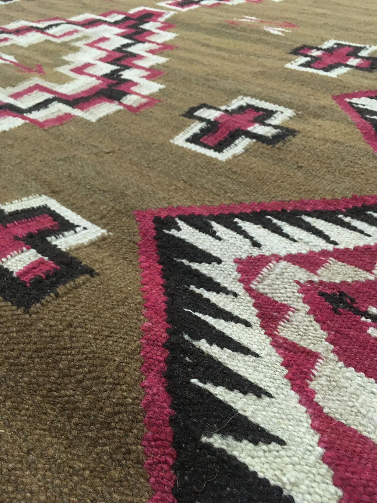 Close-up of a woven rug with a brown base and geometric Southwestern-style patterns in pink, black, and off-white. Atlantica Organics curates antique and handmade rugs, Persian, Afghan, Tibetan, Pakistani, Turkish Oushak, Kilim, Khotan, tribal and contemporary textiles for luxury interiors, homes and real estate. Our collections connect timeless craftsmanship with interior design, furniture, fine art, architecture and modern living. Explore curated rugs, collectible textiles, natural materials, cultural her
