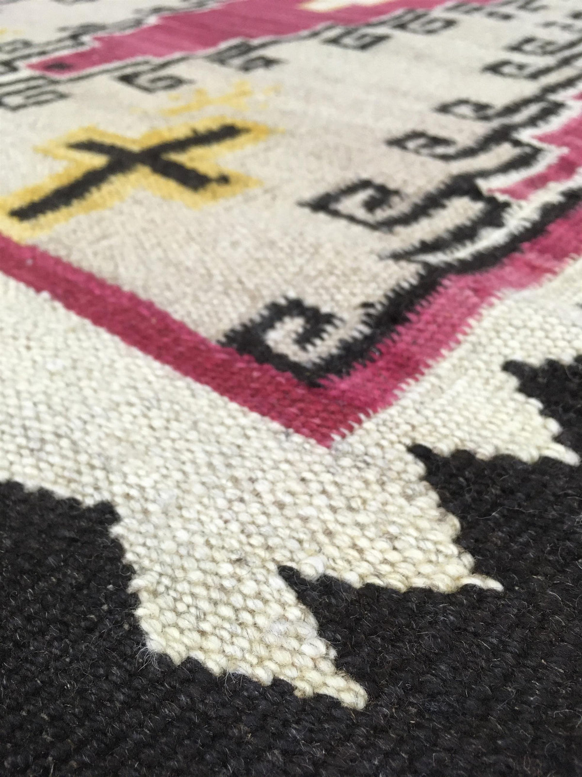 Close-up of a woven rug showing cream, black, magenta, and yellow geometric and cross-like motifs.Atlantica Organics curates antique and handmade rugs, Persian, Afghan, Tibetan, Pakistani, Turkish Oushak, Kilim, Khotan, tribal and contemporary textiles for luxury interiors, homes and real estate. Our collections connect timeless craftsmanship with interior design, furniture, fine art, architecture and modern living. Explore curated rugs, collectible textiles, natural materials, cultural heritage, sustainabl
