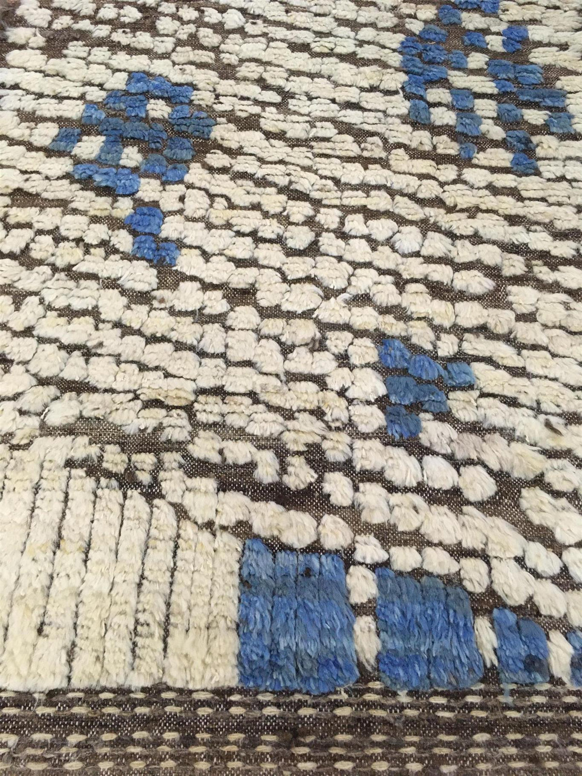 A close-up of a textured woven rug with a cream and brown base and blue geometric accents.Atlantica Organics curates antique and handmade rugs, Persian, Afghan, Tibetan, Pakistani, Turkish Oushak, Kilim, Khotan, tribal and contemporary textiles for luxury interiors, homes and real estate. Our collections connect timeless craftsmanship with interior design, furniture, fine art, architecture and modern living. Explore curated rugs, collectible textiles, natural materials, cultural heritage, sustainable design