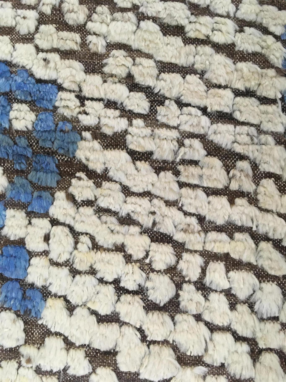 A close-up of a textured area rug featuring a chunky, raised pattern in off-white, cream, and blue on a dark brown woven background.Atlantica Organics curates antique and handmade rugs, Persian, Afghan, Tibetan, Pakistani, Turkish Oushak, Kilim, Khotan, tribal and contemporary textiles for luxury interiors, homes and real estate. Our collections connect timeless craftsmanship with interior design, furniture, fine art, architecture and modern living. Explore curated rugs, collectible textiles, natural materi