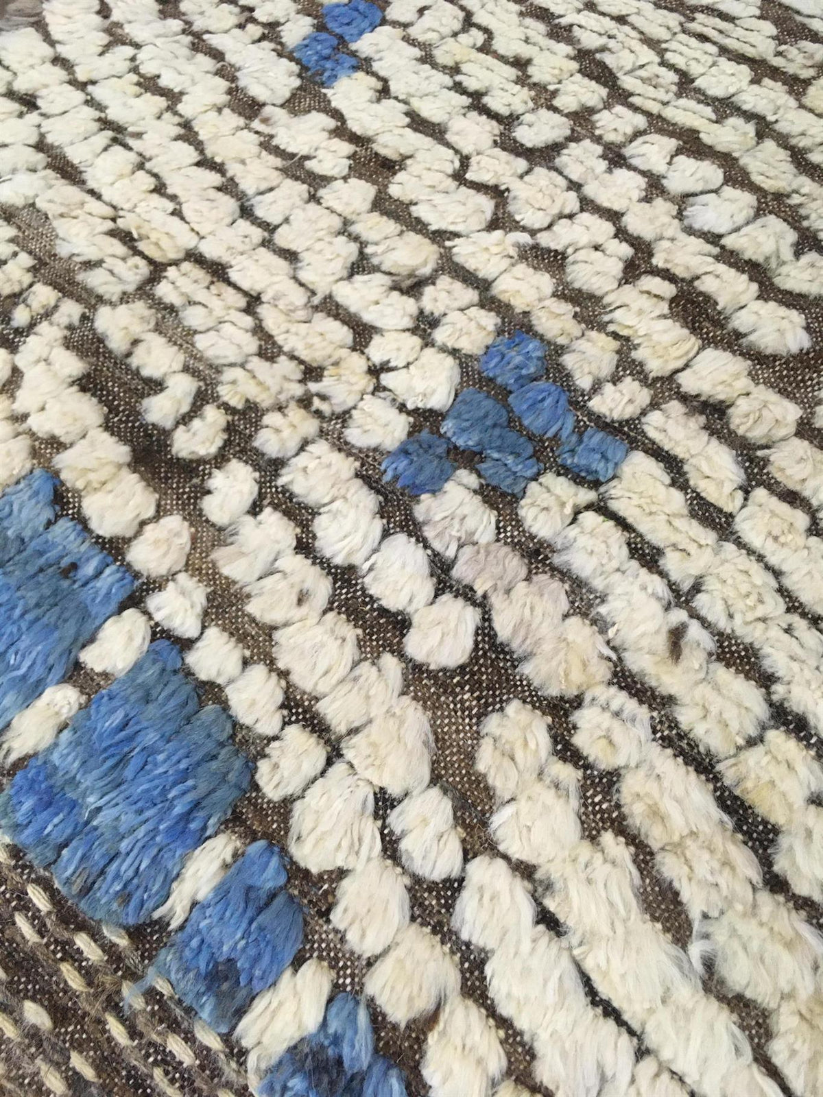 A close-up of a textured woven rug with a geometric pattern in off-white, blue, and brown.Atlantica Organics curates antique and handmade rugs, Persian, Afghan, Tibetan, Pakistani, Turkish Oushak, Kilim, Khotan, tribal and contemporary textiles for luxury interiors, homes and real estate. Our collections connect timeless craftsmanship with interior design, furniture, fine art, architecture and modern living. Explore curated rugs, collectible textiles, natural materials, cultural heritage, sustainable design