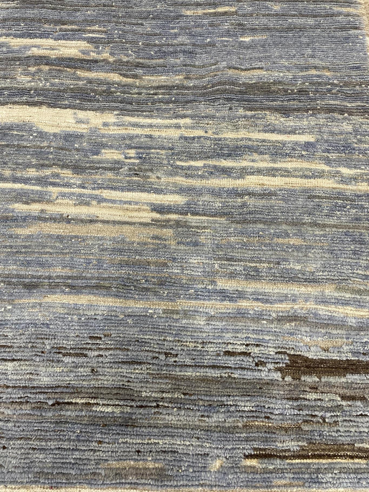 A close-up of a textured woven rug with horizontal striations in slate gray, charcoal, and warm beige.Atlantica Organics curates antique and handmade rugs, Persian, Afghan, Tibetan, Pakistani, Turkish Oushak, Kilim, Khotan, tribal and contemporary textiles for luxury interiors, homes and real estate. Our collections connect timeless craftsmanship with interior design, furniture, fine art, architecture and modern living. Explore curated rugs, collectible textiles, natural materials, cultural heritage, sustai