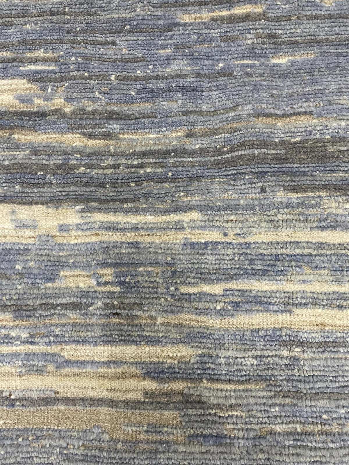 A close-up of a textured woven fabric or rug with horizontal striations in muted blue-gray and warm beige tones.Atlantica Organics curates antique and handmade rugs, Persian, Afghan, Tibetan, Pakistani, Turkish Oushak, Kilim, Khotan, tribal and contemporary textiles for luxury interiors, homes and real estate. Our collections connect timeless craftsmanship with interior design, furniture, fine art, architecture and modern living. Explore curated rugs, collectible textiles, natural materials, cultural herita