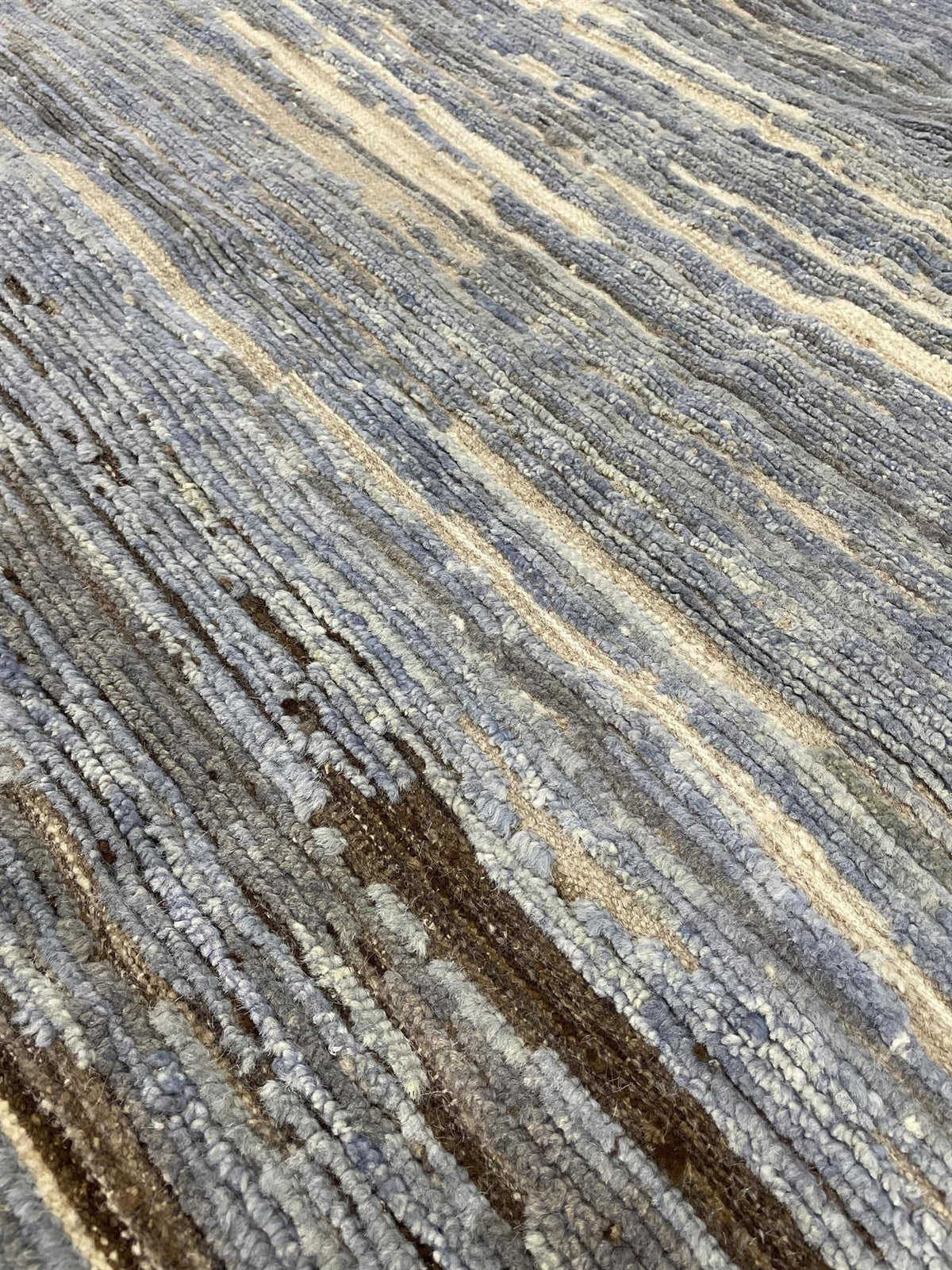 A close-up of a textured area rug with a modern linear pattern in shades of slate blue, gray, beige, and dark brown.Atlantica Organics curates antique and handmade rugs, Persian, Afghan, Tibetan, Pakistani, Turkish Oushak, Kilim, Khotan, tribal and contemporary textiles for luxury interiors, homes and real estate. Our collections connect timeless craftsmanship with interior design, furniture, fine art, architecture and modern living. Explore curated rugs, collectible textiles, natural materials, cultural he