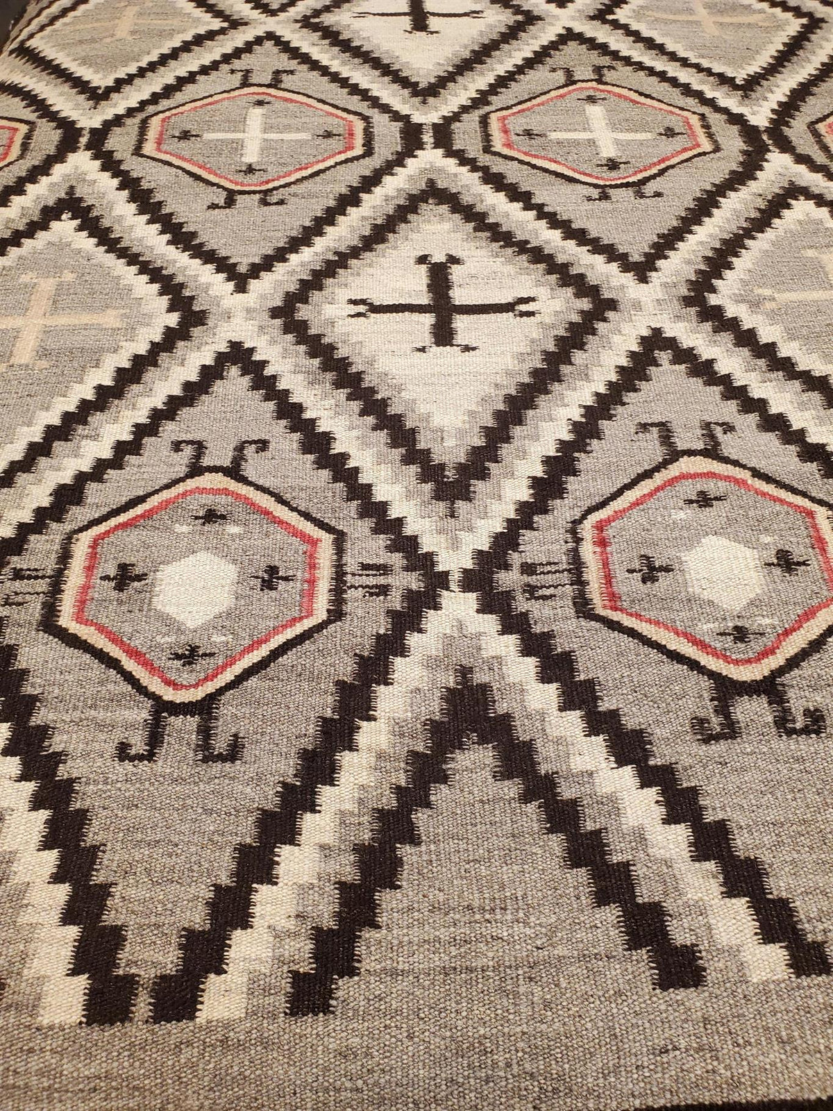 A close-up of a woven geometric rug with a Southwestern or Navajo-inspired pattern. The design features diamond and triangular motifs in charcoal gray, cream, black, and red.Atlantica Organics curates antique and handmade rugs, Persian, Afghan, Tibetan, Pakistani, Turkish Oushak, Kilim, Khotan, tribal and contemporary textiles for luxury interiors, homes and real estate. Our collections connect timeless craftsmanship with interior design, furniture, fine art, architecture and modern living. Explore curated 