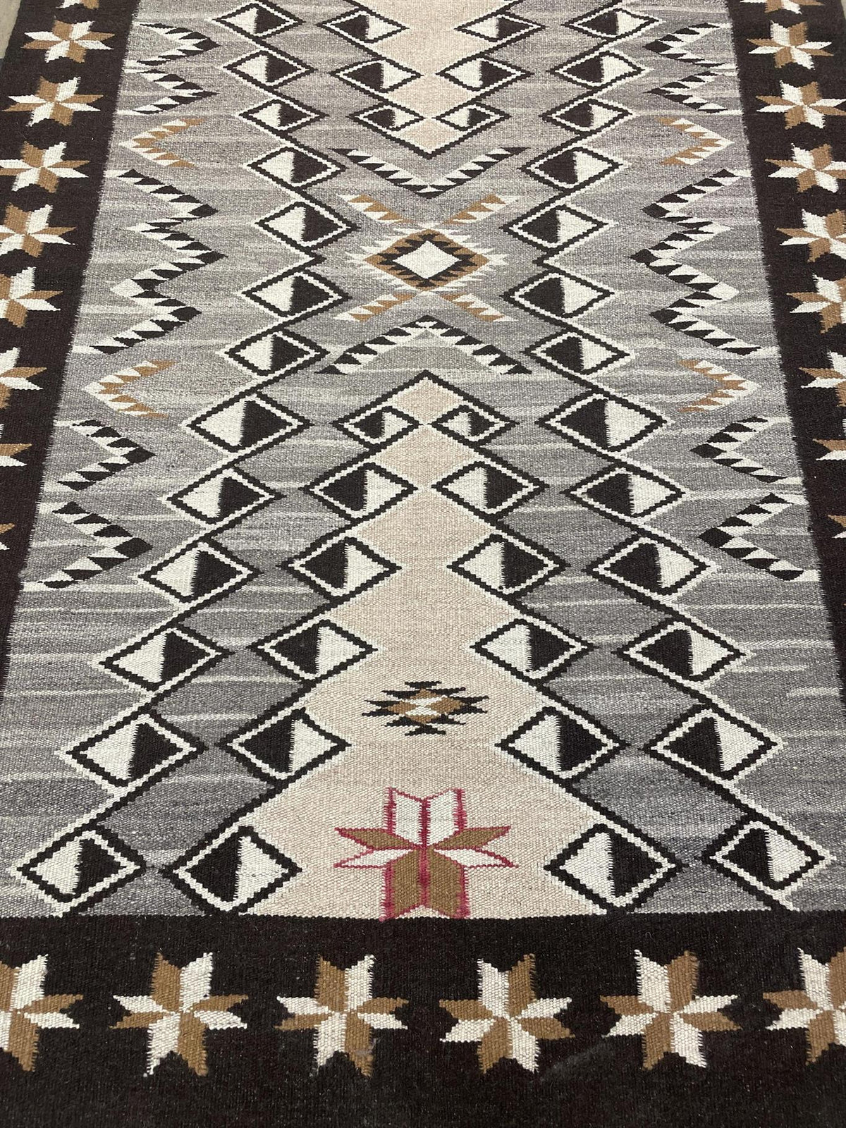 A close-up of a woven geometric rug with a Southwestern or Navajo-inspired pattern. The design features a central field of black, white, and gray diamonds and triangles, bordered by a cream and brown star pattern on a dark charcoal base. Atlantica Organics curates antique and handmade rugs, Persian, Afghan, Tibetan, Pakistani, Turkish Oushak, Kilim, Khotan, tribal and contemporary textiles for luxury interiors, homes and real estate. Our collections connect timeless craftsmanship with interior design, furni