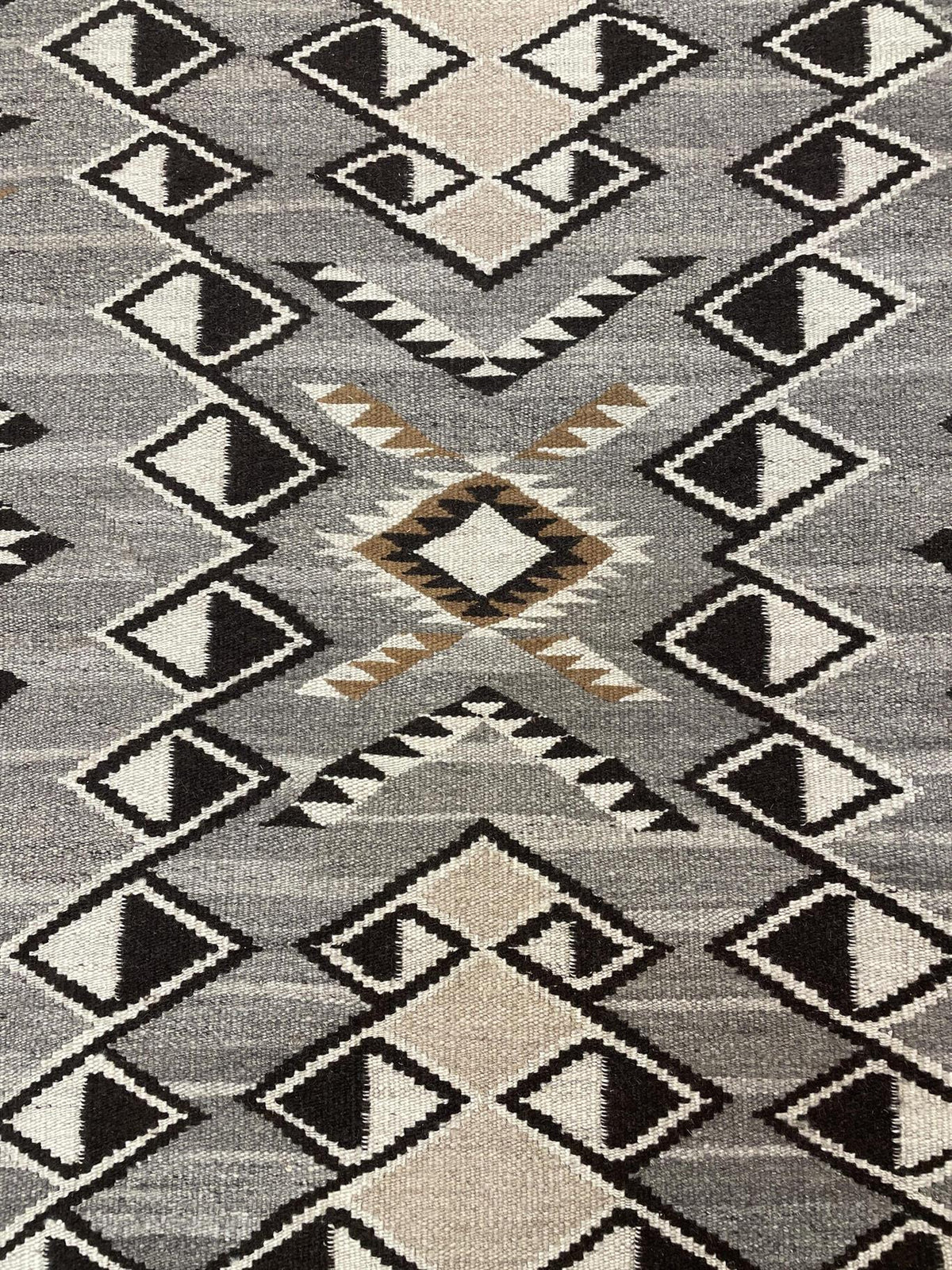 A close-up of a woven rug with a Southwestern geometric pattern in gray, beige, black, and brown. Atlantica Organics curates antique and handmade rugs, Persian, Afghan, Tibetan, Pakistani, Turkish Oushak, Kilim, Khotan, tribal and contemporary textiles for luxury interiors, homes and real estate. Our collections connect timeless craftsmanship with interior design, furniture, fine art, architecture and modern living. Explore curated rugs, collectible textiles, natural materials, cultural heritage, sustainabl