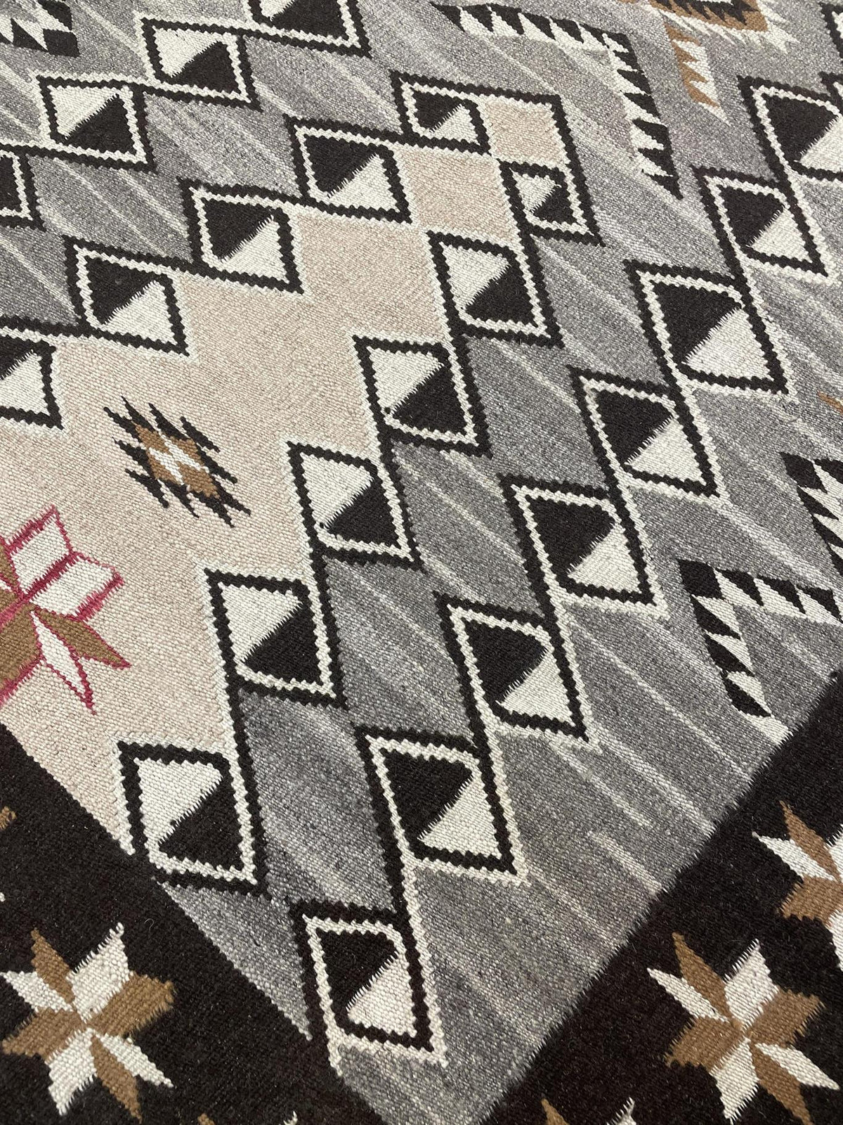 Close-up of a woven rug with a Southwestern geometric pattern in gray, black, beige, white, and brown. Atlantica Organics curates antique and handmade rugs, Persian, Afghan, Tibetan, Pakistani, Turkish Oushak, Kilim, Khotan, tribal and contemporary textiles for luxury interiors, homes and real estate. Our collections connect timeless craftsmanship with interior design, furniture, fine art, architecture and modern living. Explore curated rugs, collectible textiles, natural materials, cultural heritage, susta