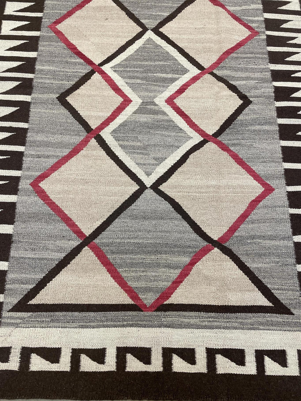 A close-up of a woven rug with a geometric pattern featuring large diamonds and triangles in beige, gray, black, and muted red, bordered by black and white zigzag patterns.Atlantica Organics curates antique and handmade rugs, Persian, Afghan, Tibetan, Pakistani, Turkish Oushak, Kilim, Khotan, tribal and contemporary textiles for luxury interiors, homes and real estate. Our collections connect timeless craftsmanship with interior design, furniture, fine art, architecture and modern living. Explore curated ru