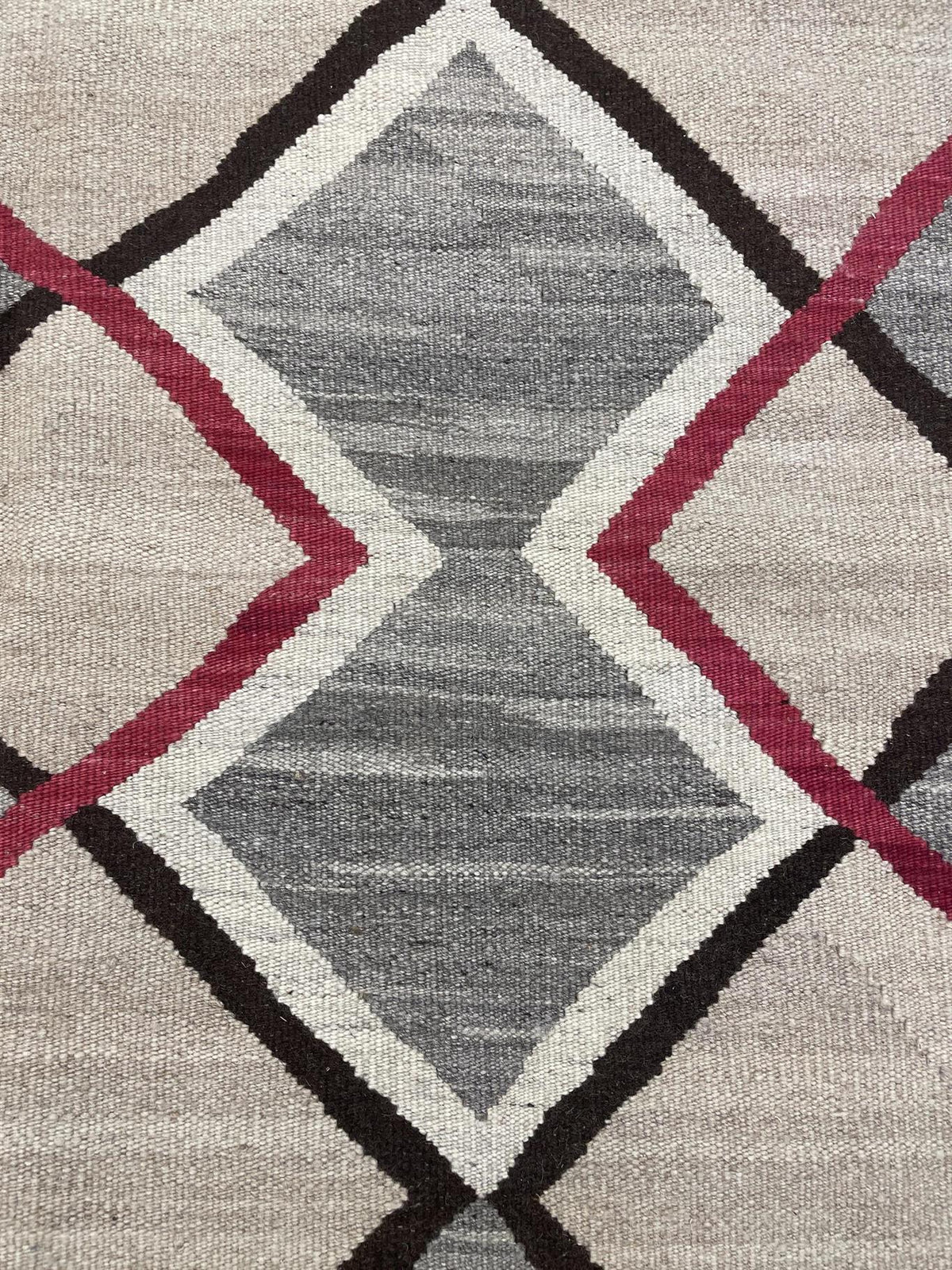 A close-up of a woven textile with a geometric diamond pattern in gray, beige, white, black, and muted red.Atlantica Organics curates antique and handmade rugs, Persian, Afghan, Tibetan, Pakistani, Turkish Oushak, Kilim, Khotan, tribal and contemporary textiles for luxury interiors, homes and real estate. Our collections connect timeless craftsmanship with interior design, furniture, fine art, architecture and modern living. Explore curated rugs, collectible textiles, natural materials, cultural heritage, s