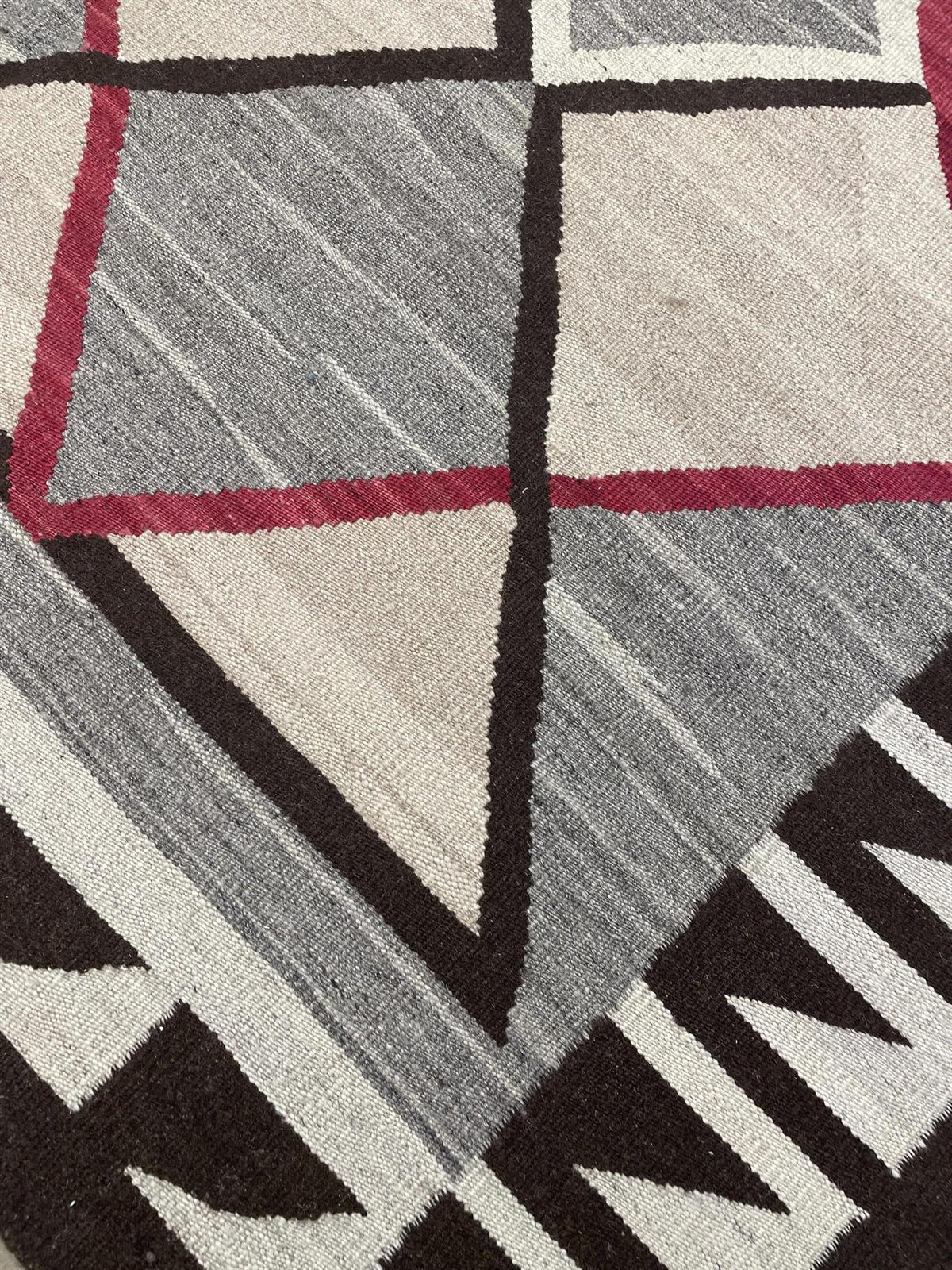 Close-up of a geometric patterned rug with gray, beige, black, and red sections, showing the woven texture and design details.Atlantica Organics curates antique and handmade rugs, Persian, Afghan, Tibetan, Pakistani, Turkish Oushak, Kilim, Khotan, tribal and contemporary textiles for luxury interiors, homes and real estate. Our collections connect timeless craftsmanship with interior design, furniture, fine art, architecture and modern living. Explore curated rugs, collectible textiles, natural materials, c