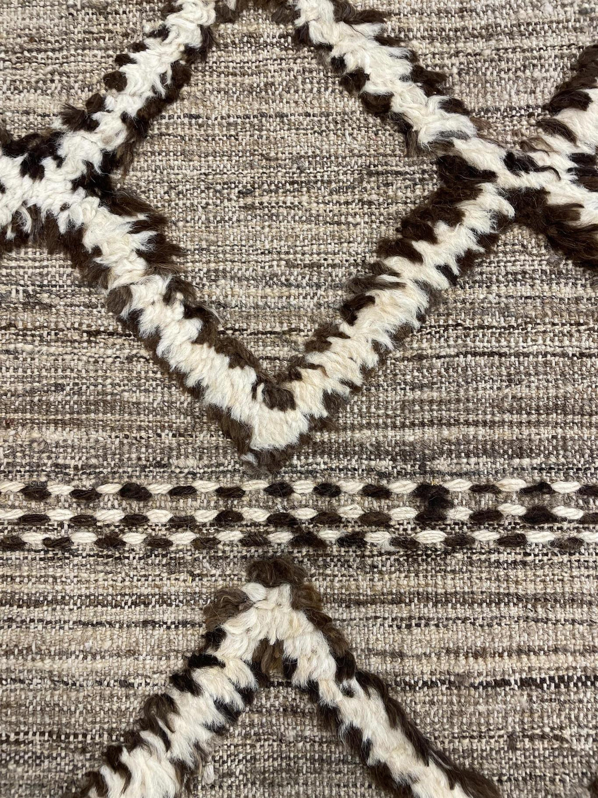 Close-up of a woven rug or textile with a geometric diamond pattern in cream and dark brown on a taupe-beige background.Atlantica Organics curates antique and handmade rugs, Persian, Afghan, Tibetan, Pakistani, Turkish Oushak, Kilim, Khotan, tribal and contemporary textiles for luxury interiors, homes and real estate. Our collections connect timeless craftsmanship with interior design, furniture, fine art, architecture and modern living. Explore curated rugs, collectible textiles, natural materials, cultura