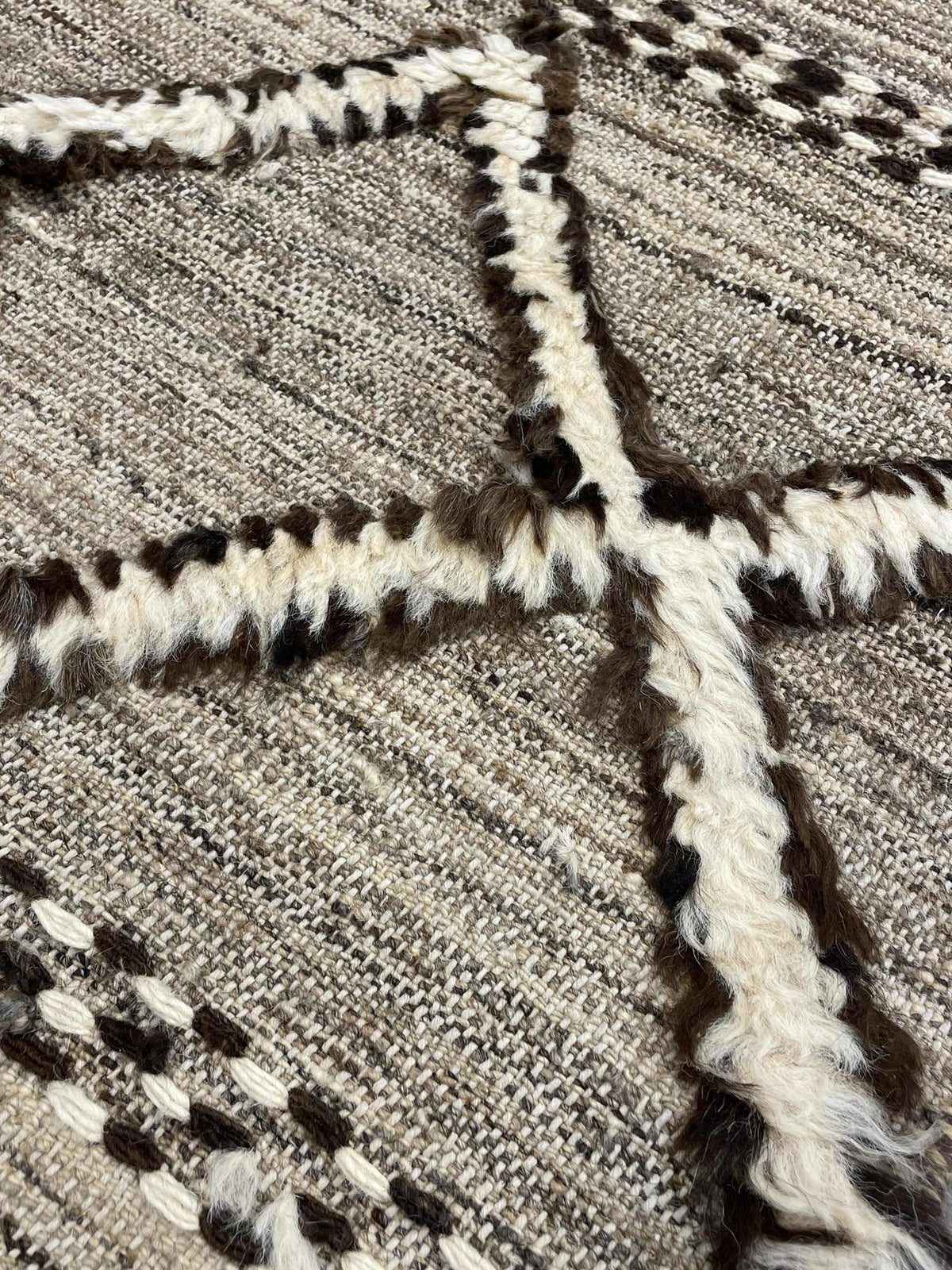 Close-up of a woven rug with a textured geometric pattern in beige, brown, and off-white.Atlantica Organics curates antique and handmade rugs, Persian, Afghan, Tibetan, Pakistani, Turkish Oushak, Kilim, Khotan, tribal and contemporary textiles for luxury interiors, homes and real estate. Our collections connect timeless craftsmanship with interior design, furniture, fine art, architecture and modern living. Explore curated rugs, collectible textiles, natural materials, cultural heritage, sustainable design 