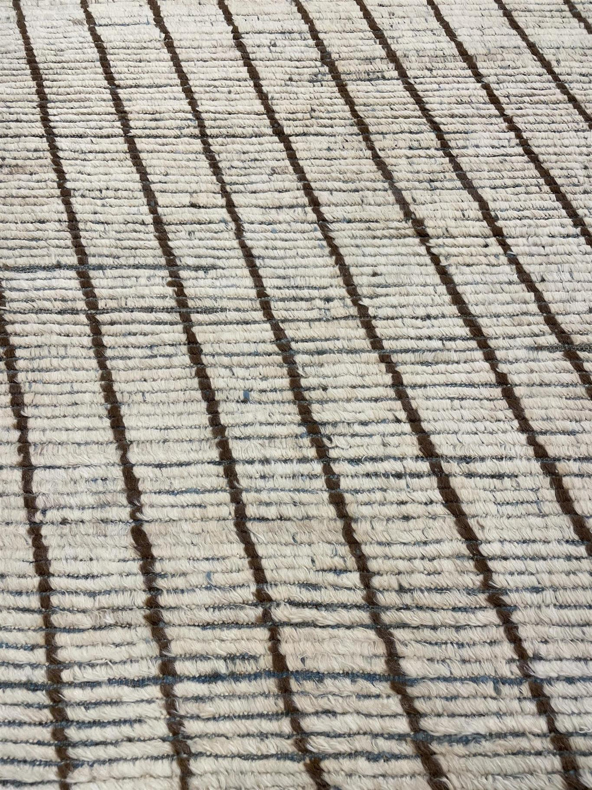 Close-up of a textured rug with a cream base and dark brown vertical stripes, accented by thin blue horizontal lines. Atlantica Organics curates antique and handmade rugs, Persian, Afghan, Tibetan, Pakistani, Turkish Oushak, Kilim, Khotan, tribal and contemporary textiles for luxury interiors, homes and real estate. Our collections connect timeless craftsmanship with interior design, furniture, fine art, architecture and modern living. Explore curated rugs, collectible textiles, natural materials, cultural 