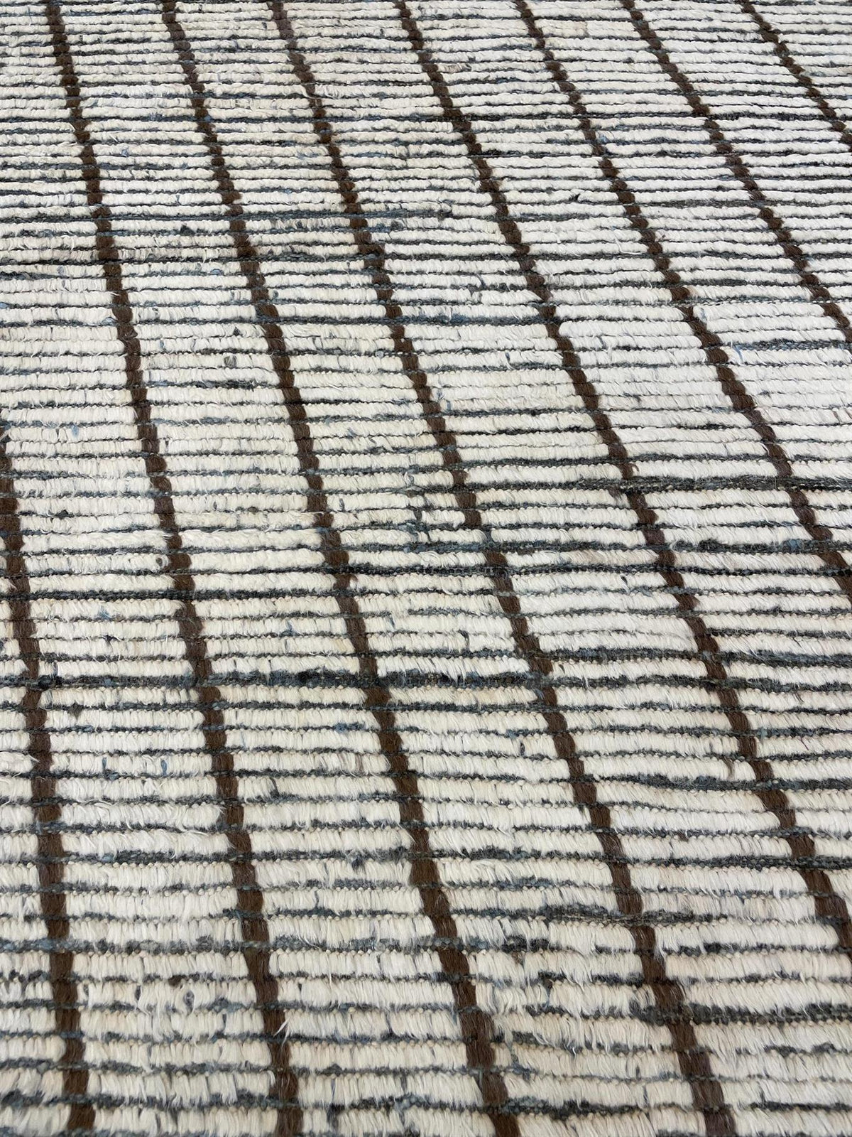 Close-up of a textured woven rug with a cream base and dark brown vertical stripe pattern. Atlantica Organics curates antique and handmade rugs, Persian, Afghan, Tibetan, Pakistani, Turkish Oushak, Kilim, Khotan, tribal and contemporary textiles for luxury interiors, homes and real estate. Our collections connect timeless craftsmanship with interior design, furniture, fine art, architecture and modern living. Explore curated rugs, collectible textiles, natural materials, cultural heritage, sustainable desig