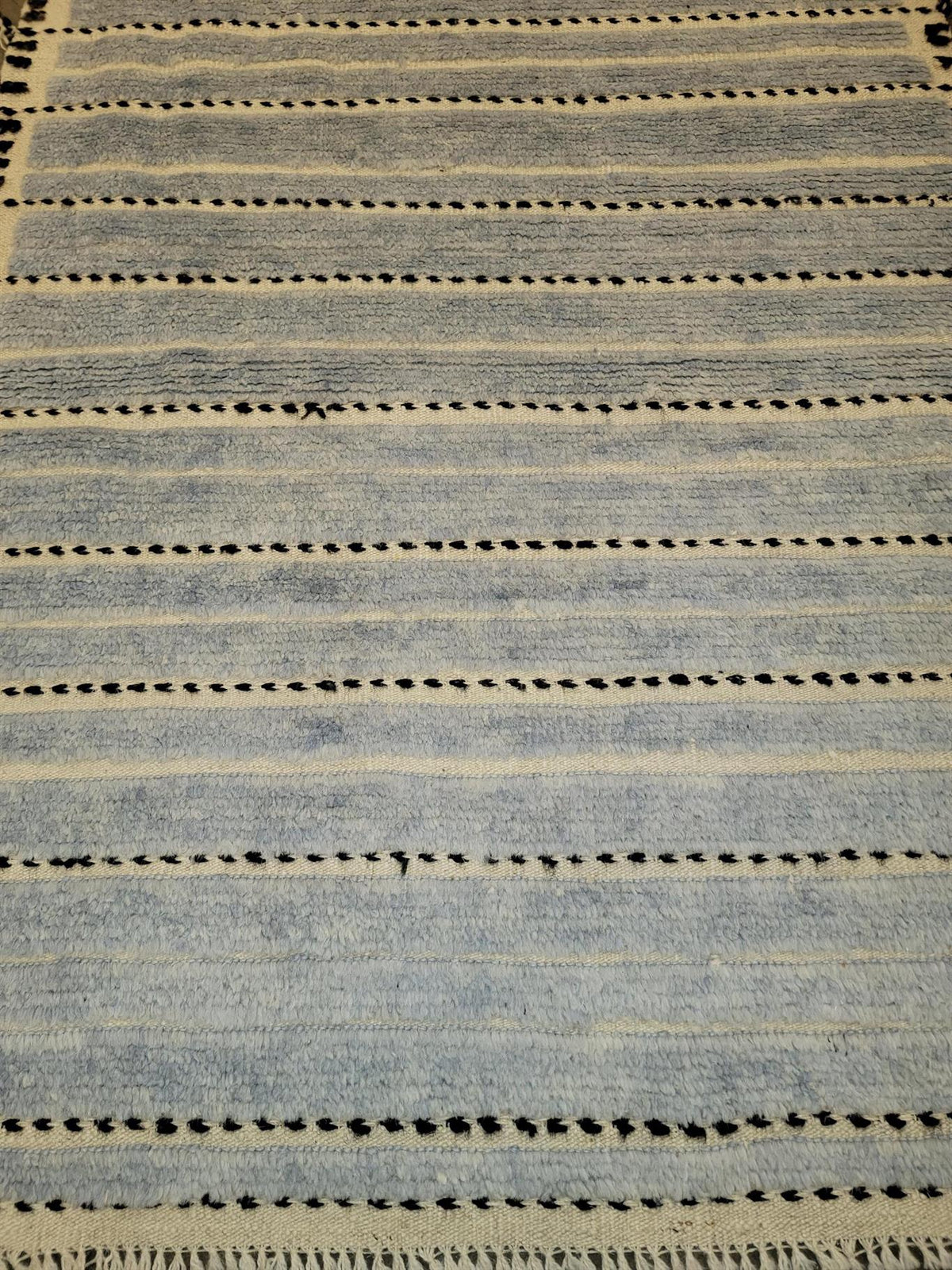 A close-up of a woven rug with horizontal stripes in light blue, beige, and black.Atlantica Organics curates antique and handmade rugs, Persian, Afghan, Tibetan, Pakistani, Turkish Oushak, Kilim, Khotan, tribal and contemporary textiles for luxury interiors, homes and real estate. Our collections connect timeless craftsmanship with interior design, furniture, fine art, architecture and modern living. Explore curated rugs, collectible textiles, natural materials, cultural heritage, sustainable design and dis