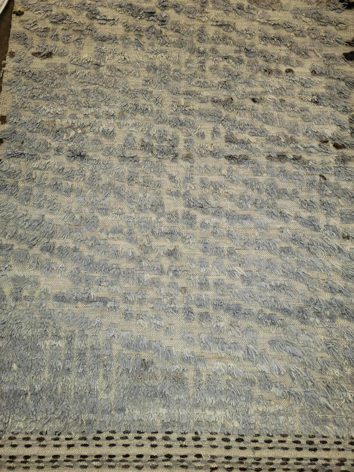 A close-up of a textured gray and beige woven rug or mat with a perforated border along the bottom edge. Atlantica Organics curates antique and handmade rugs, Persian, Afghan, Tibetan, Pakistani, Turkish Oushak, Kilim, Khotan, tribal and contemporary textiles for luxury interiors, homes and real estate. Our collections connect timeless craftsmanship with interior design, furniture, fine art, architecture and modern living. Explore curated rugs, collectible textiles, natural materials, cultural heritage, sus