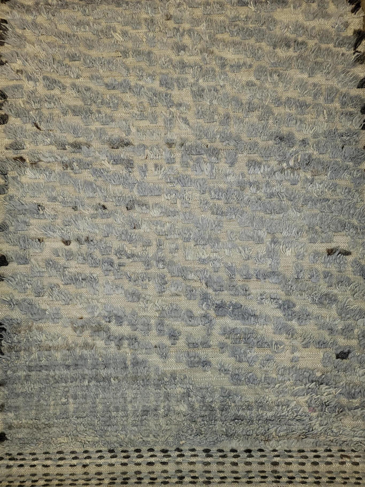 A close-up of a textured woven rug or textile in muted blue-gray and beige tones, showing the fabric weave and a dark dotted border along the bottom edge. Atlantica Organics curates antique and handmade rugs, Persian, Afghan, Tibetan, Pakistani, Turkish Oushak, Kilim, Khotan, tribal and contemporary textiles for luxury interiors, homes and real estate. Our collections connect timeless craftsmanship with interior design, furniture, fine art, architecture and modern living. Explore curated rugs, collectible t
