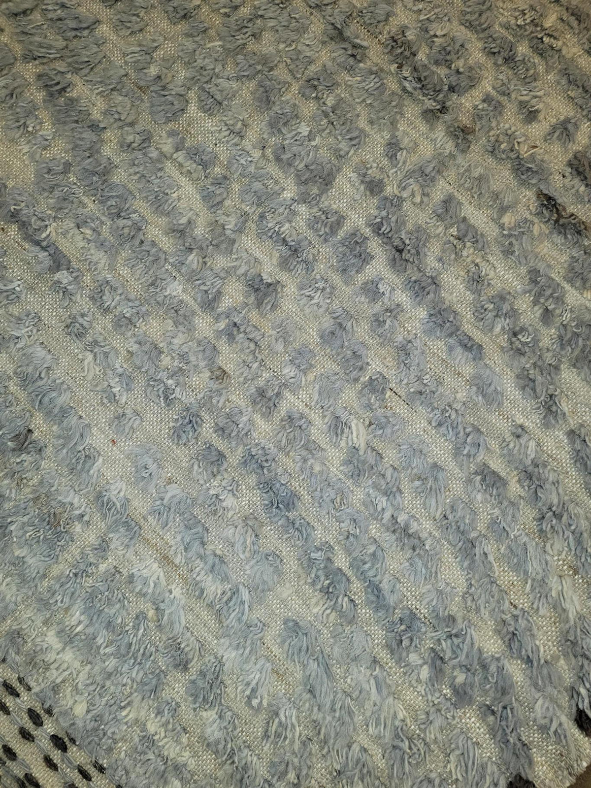A close-up of a textured rug with a shaggy, looped pile in a mix of gray and beige tones, arranged in diagonal ribbed stripes. Atlantica Organics curates antique and handmade rugs, Persian, Afghan, Tibetan, Pakistani, Turkish Oushak, Kilim, Khotan, tribal and contemporary textiles for luxury interiors, homes and real estate. Our collections connect timeless craftsmanship with interior design, furniture, fine art, architecture and modern living. Explore curated rugs, collectible textiles, natural materials, 