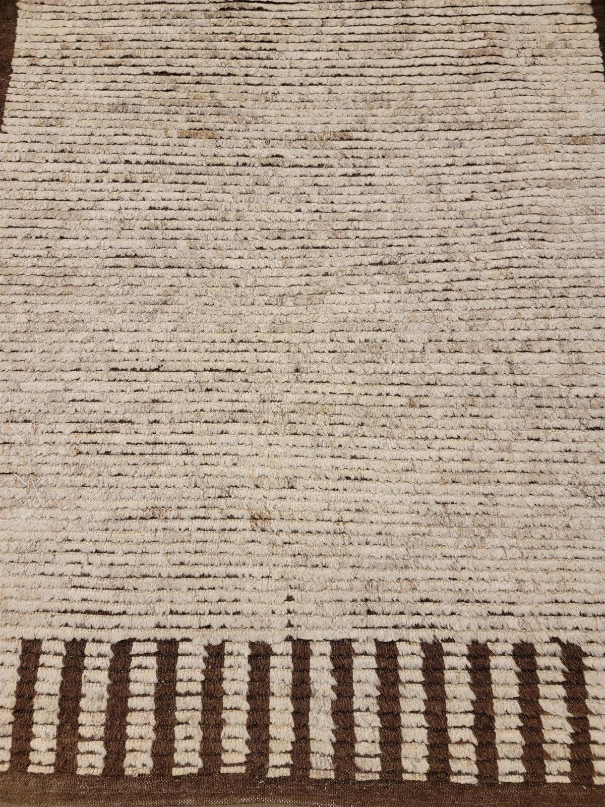 A close-up of a woven rug showing a textured cream and brown striped pattern with a decorative border at the bottom.Atlantica Organics curates antique and handmade rugs, Persian, Afghan, Tibetan, Pakistani, Turkish Oushak, Kilim, Khotan, tribal and contemporary textiles for luxury interiors, homes and real estate. Our collections connect timeless craftsmanship with interior design, furniture, fine art, architecture and modern living. Explore curated rugs, collectible textiles, natural materials, cultural he