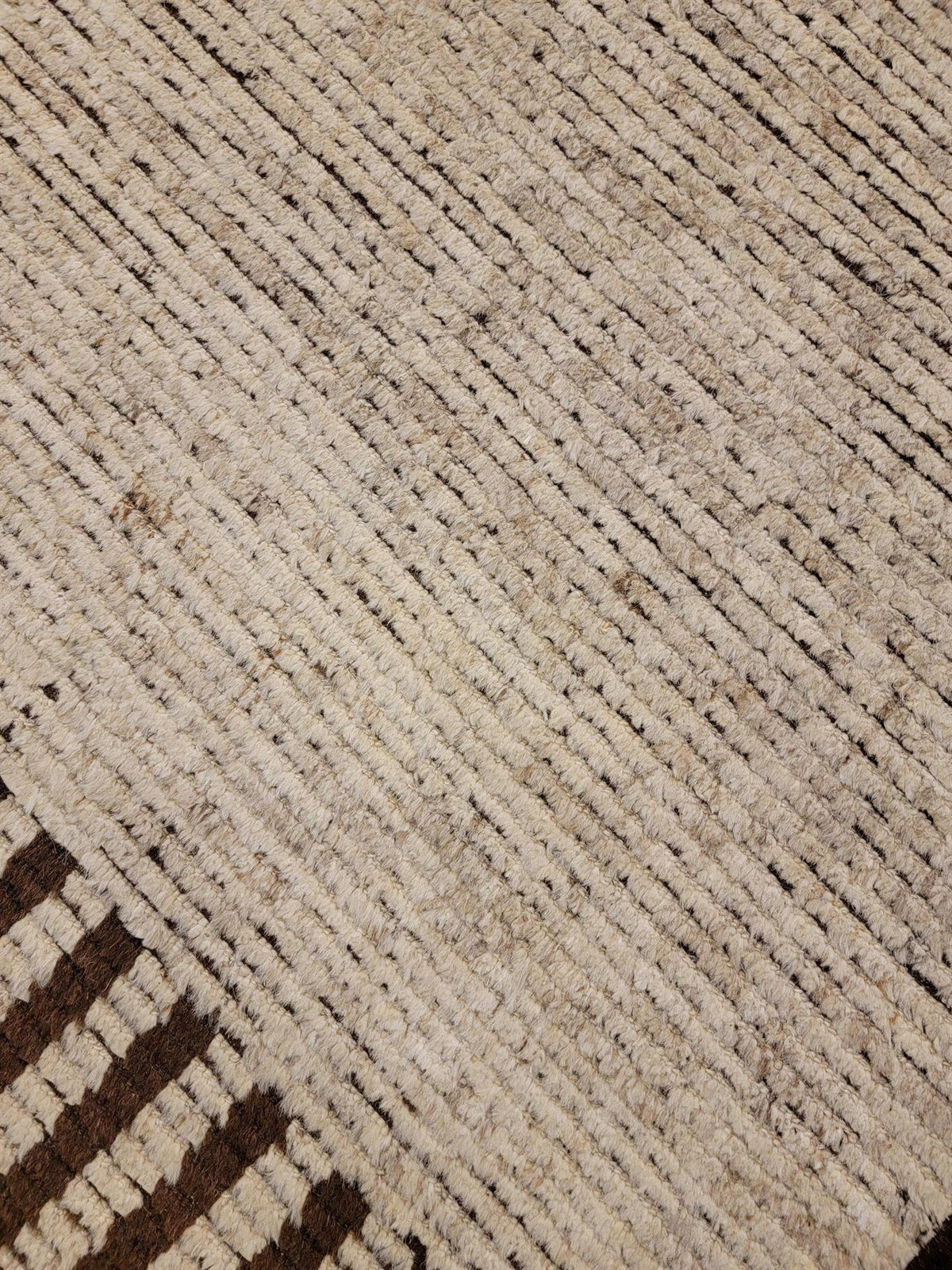 Close-up of a woven rug or textile showing a cream and beige base with dark brown linear patterning and a small section of dark brown striped border in the lower left corner.Atlantica Organics curates antique and handmade rugs, Persian, Afghan, Tibetan, Pakistani, Turkish Oushak, Kilim, Khotan, tribal and contemporary textiles for luxury interiors, homes and real estate. Our collections connect timeless craftsmanship with interior design, furniture, fine art, architecture and modern living. Explore curated 