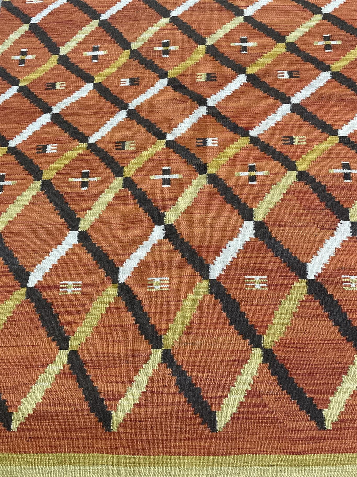 A close-up of a woven textile or rug showing a geometric diamond pattern in rust orange, black, white, and olive green.Atlantica Organics curates antique and handmade rugs, Persian, Afghan, Tibetan, Pakistani, Turkish Oushak, Kilim, Khotan, tribal and contemporary textiles for luxury interiors, homes and real estate. Our collections connect timeless craftsmanship with interior design, furniture, fine art, architecture and modern living. Explore curated rugs, collectible textiles, natural materials, cultural