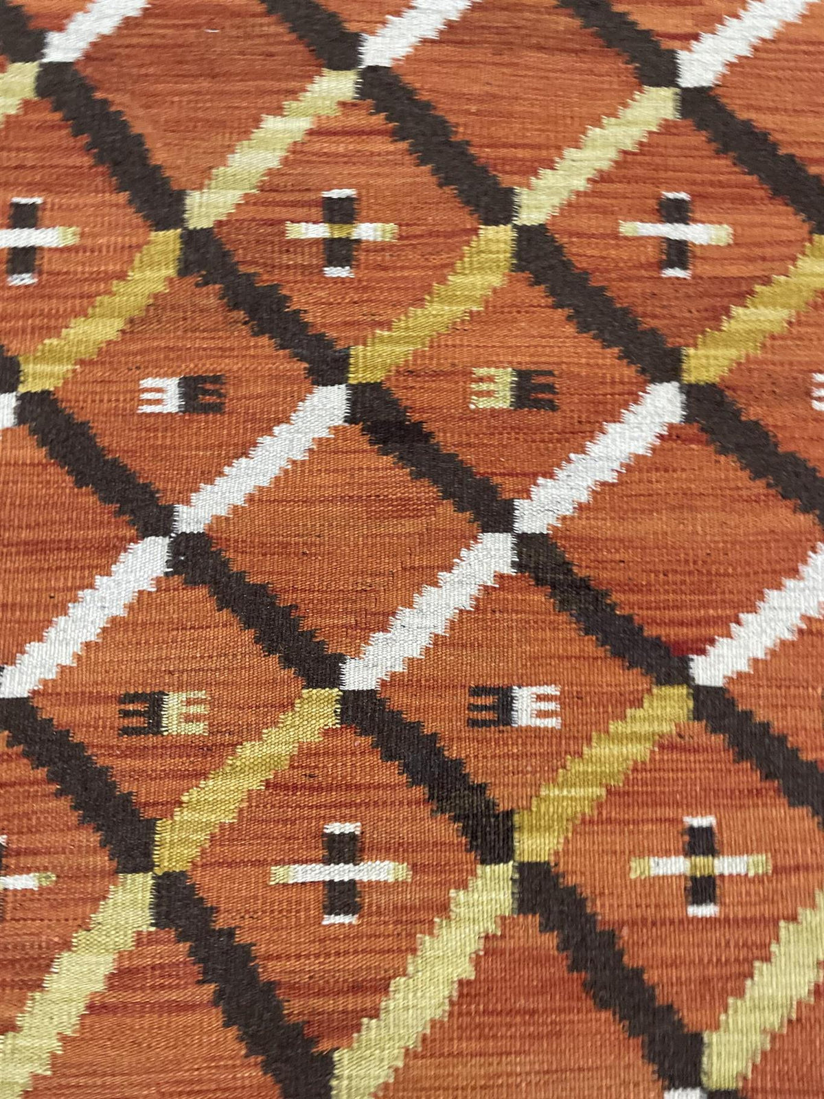 A close-up of a woven textile with a geometric diamond pattern in rust orange, black, olive green, white, and tan.Atlantica Organics curates antique and handmade rugs, Persian, Afghan, Tibetan, Pakistani, Turkish Oushak, Kilim, Khotan, tribal and contemporary textiles for luxury interiors, homes and real estate. Our collections connect timeless craftsmanship with interior design, furniture, fine art, architecture and modern living. Explore curated rugs, collectible textiles, natural materials, cultural heri