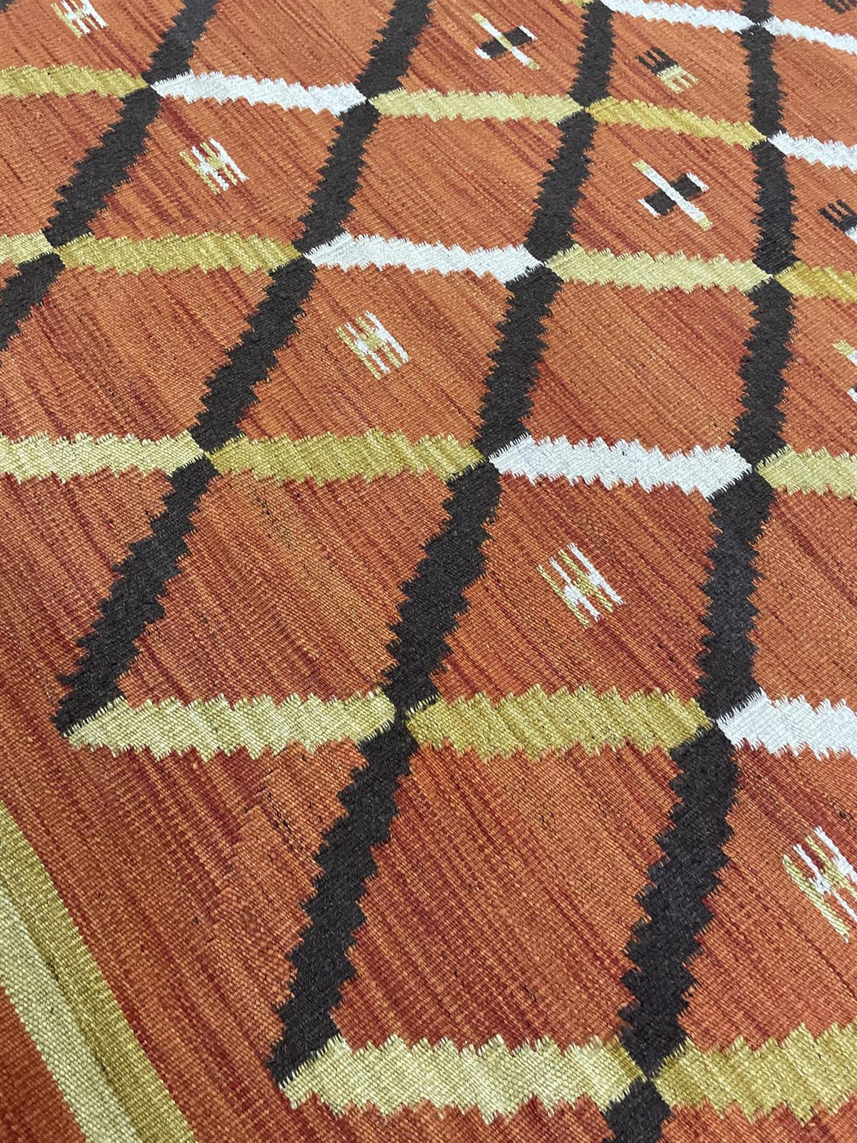 A close-up of a woven textile or rug showing a geometric diamond pattern in rust orange, black, olive green, white, and tan.Atlantica Organics curates antique and handmade rugs, Persian, Afghan, Tibetan, Pakistani, Turkish Oushak, Kilim, Khotan, tribal and contemporary textiles for luxury interiors, homes and real estate. Our collections connect timeless craftsmanship with interior design, furniture, fine art, architecture and modern living. Explore curated rugs, collectible textiles, natural materials, cul