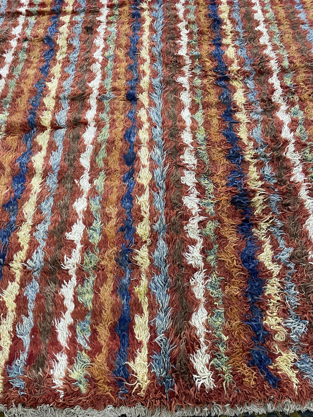 A close-up of a shaggy area rug with vertical stripes in rust red, brown, cream, blue, and tan. Atlantica Organics curates antique and handmade rugs, Persian, Afghan, Tibetan, Pakistani, Turkish Oushak, Kilim, Khotan, tribal and contemporary textiles for luxury interiors, homes and real estate. Our collections connect timeless craftsmanship with interior design, furniture, fine art, architecture and modern living. Explore curated rugs, collectible textiles, natural materials, cultural heritage, sustainable 