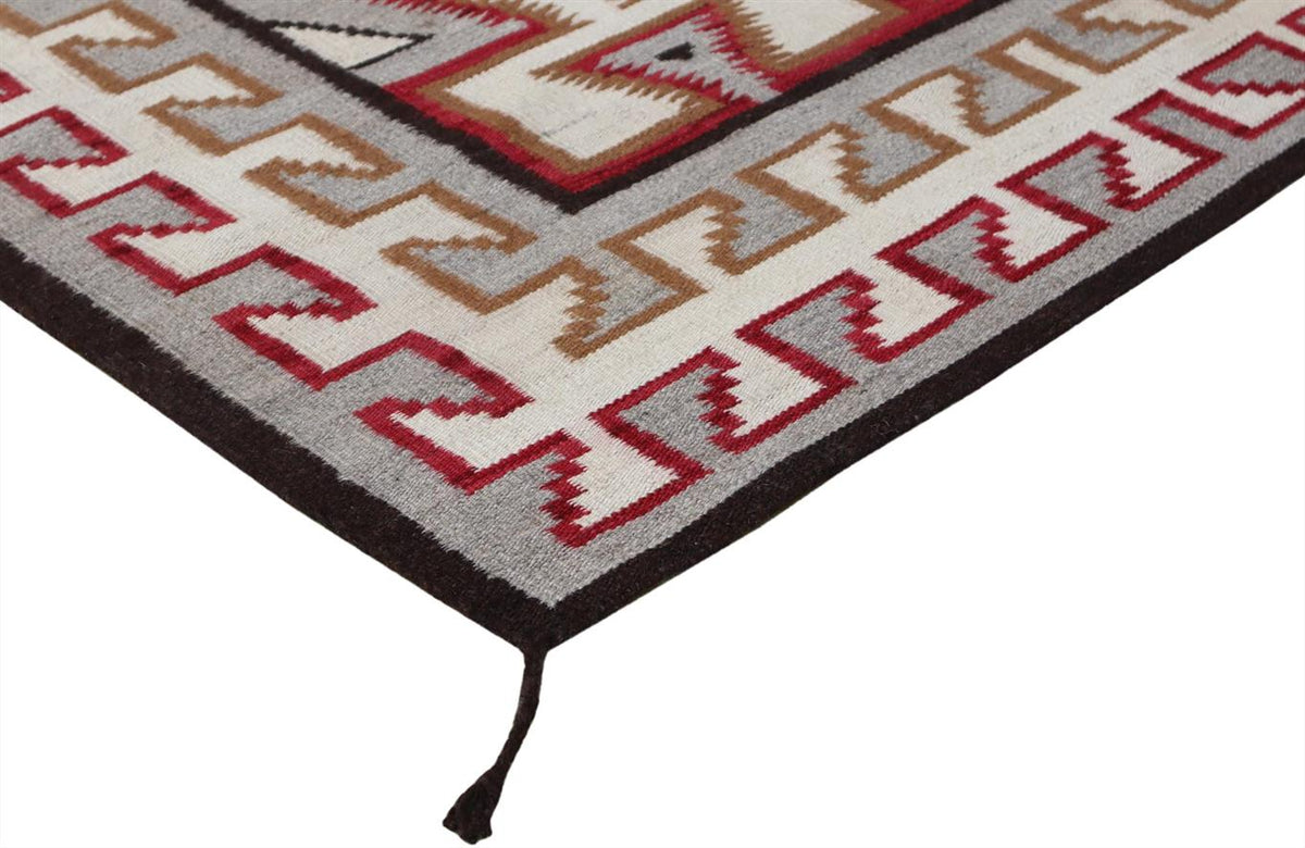 A close-up of a woven rug corner showing a geometric Southwestern-style pattern in red, brown, gray, and cream with a dark border and tassel.