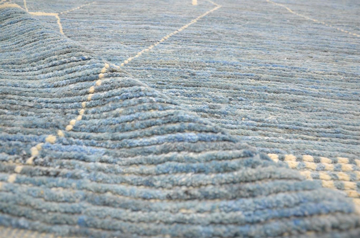 A close-up of a textured blue rug with horizontal ribbed stripes and light beige geometric line accents. Atlantica Organics curates antique and handmade rugs, Persian, Afghan, Tibetan, Pakistani, Turkish Oushak, Kilim, Khotan, tribal and contemporary textiles for luxury interiors, homes and real estate. Our collections connect timeless craftsmanship with interior design, furniture, fine art, architecture and modern living. Explore curated rugs, collectible textiles, natural materials, cultural heritage, sus