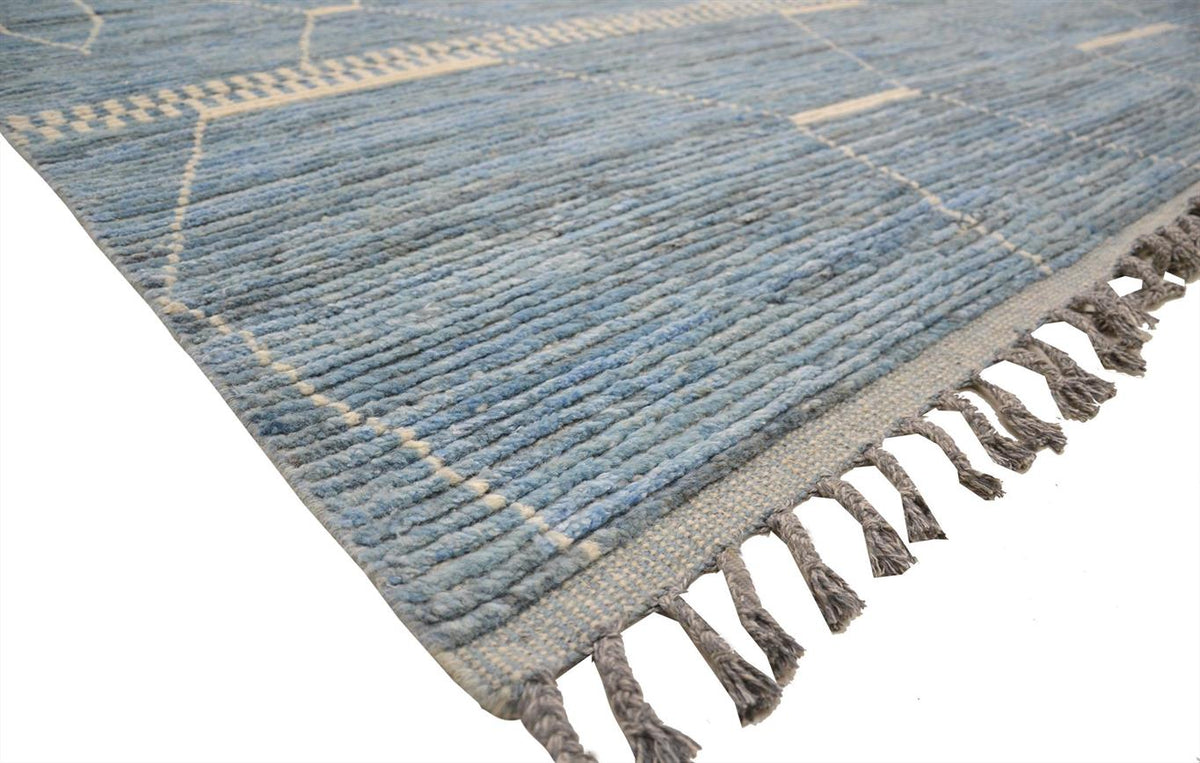 A close-up of a blue woven rug with a textured geometric pattern and dark gray fringe tassels, shown on a white background. Atlantica Organics curates antique and handmade rugs, Persian, Afghan, Tibetan, Pakistani, Turkish Oushak, Kilim, Khotan, tribal and contemporary textiles for luxury interiors, homes and real estate. Our collections connect timeless craftsmanship with interior design, furniture, fine art, architecture and modern living. Explore curated rugs, collectible textiles, natural materials, cul