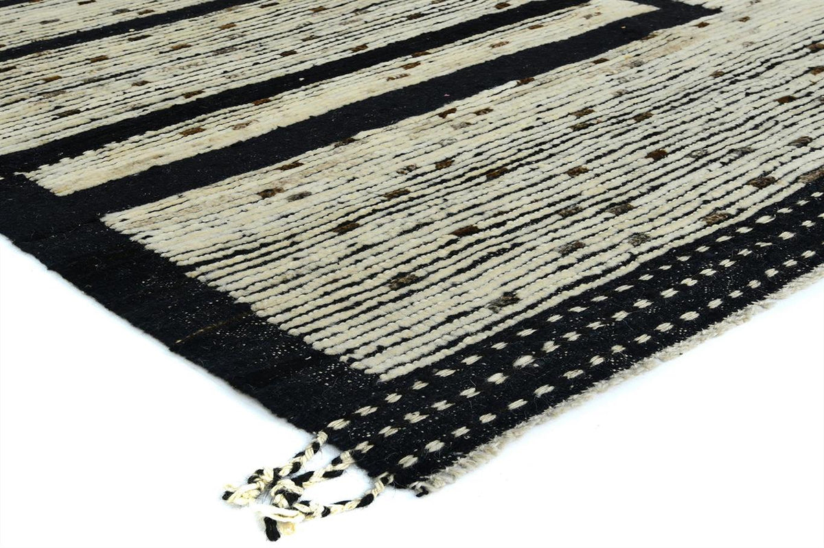 A close-up of a woven rug corner showing a black and cream striped pattern with small brown dotted accents and a fringed edge on a white background. Atlantica Organics curates antique and handmade rugs, Persian, Afghan, Tibetan, Pakistani, Turkish Oushak, Kilim, Khotan, tribal and contemporary textiles for luxury interiors, homes and real estate. Our collections connect timeless craftsmanship with interior design, furniture, fine art, architecture and modern living. Explore curated rugs, collectible textile