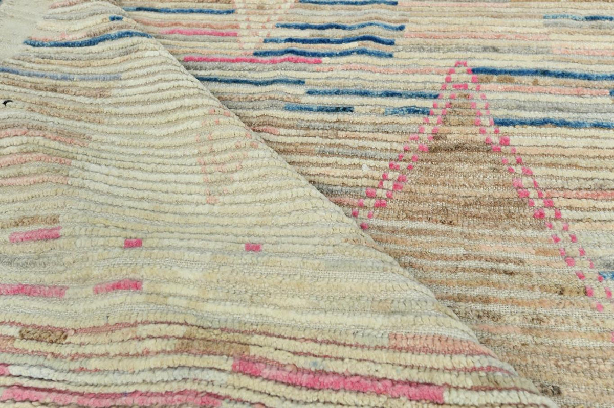 A close-up of a textured woven rug with horizontal stripes in beige, cream, brown, blue, and pink, with a folded corner revealing the pile and weave. Atlantica Organics curates antique and handmade rugs, Persian, Afghan, Tibetan, Pakistani, Turkish Oushak, Kilim, Khotan, tribal and contemporary textiles for luxury interiors, homes and real estate. Our collections connect timeless craftsmanship with interior design, furniture, fine art, architecture and modern living. Explore curated rugs, collectible textil