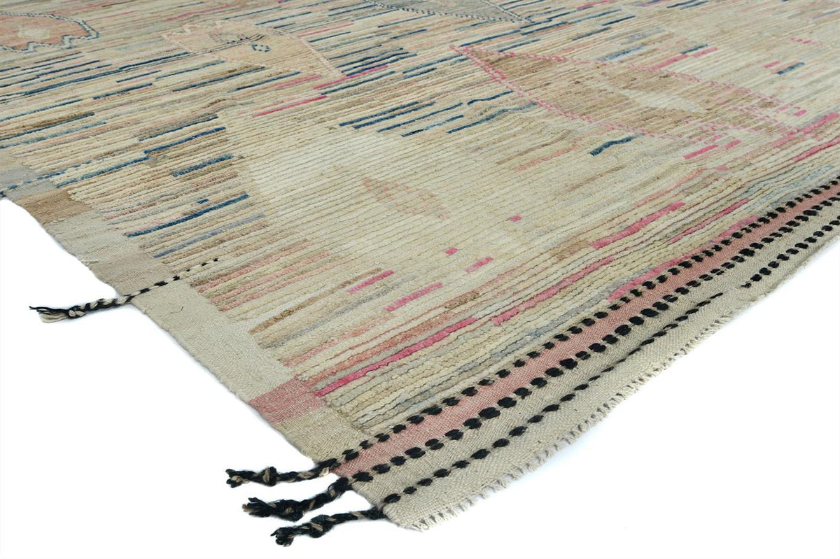 A close-up of a woven area rug with a textured, abstract pattern in beige, pink, blue, and black, featuring black tassels on one end. Atlantica Organics curates antique and handmade rugs, Persian, Afghan, Tibetan, Pakistani, Turkish Oushak, Kilim, Khotan, tribal and contemporary textiles for luxury interiors, homes and real estate. Our collections connect timeless craftsmanship with interior design, furniture, fine art, architecture and modern living. Explore curated rugs, collectible textiles, natural mate