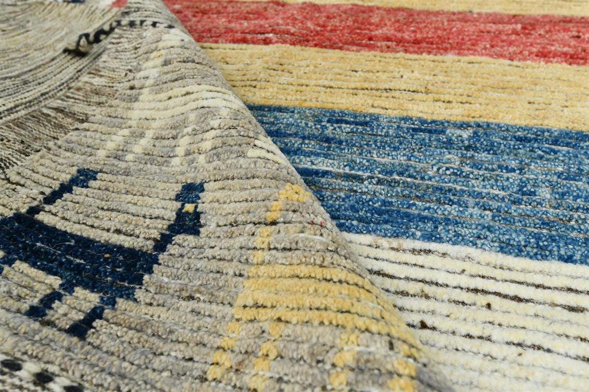 Close-up of a textured woven rug with a folded corner revealing a multicolor striped pattern in red, yellow, blue, and cream. Atlantica Organics curates antique and handmade rugs, Persian, Afghan, Tibetan, Pakistani, Turkish Oushak, Kilim, Khotan, tribal and contemporary textiles for luxury interiors, homes and real estate. Our collections connect timeless craftsmanship with interior design, furniture, fine art, architecture and modern living. Explore curated rugs, collectible textiles, natural materials, c