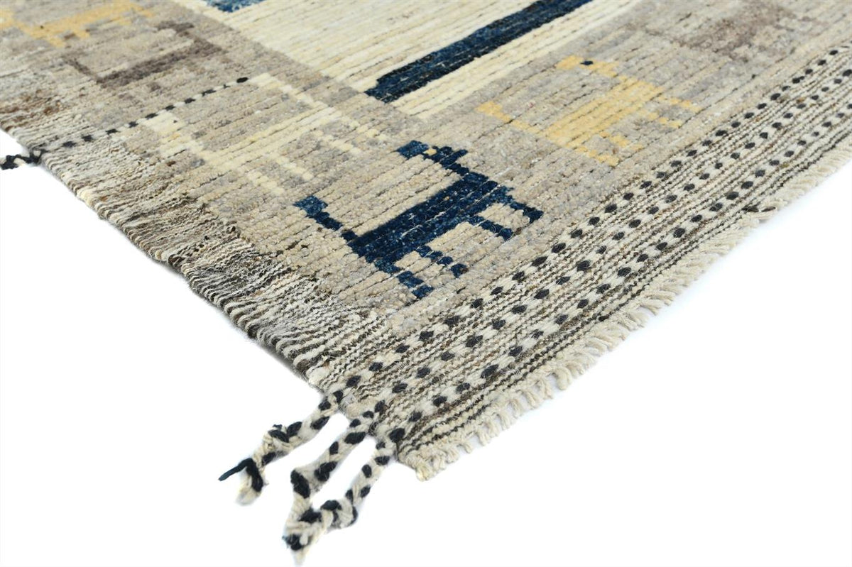 A close-up of a woven area rug with a textured, hand-knotted appearance. The rug features a geometric pattern in beige, cream, and dark navy blue, with a fringed edge and tassels visible in the foreground, set against a plain white background. Atlantica Organics curates antique and handmade rugs, Persian, Afghan, Tibetan, Pakistani, Turkish Oushak, Kilim, Khotan, tribal and contemporary textiles for luxury interiors, homes and real estate. Our collections connect timeless craftsmanship with interior design,