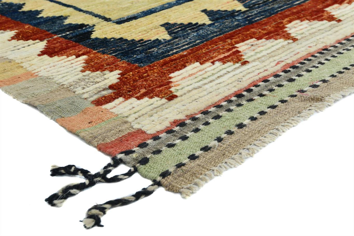 A close-up of a woven rug corner showing geometric patterns in cream, rust orange, navy blue, sage green, and black, with fringed tassels on a white background. Atlantica Organics curates antique and handmade rugs, Persian, Afghan, Tibetan, Pakistani, Turkish Oushak, Kilim, Khotan, tribal and contemporary textiles for luxury interiors, homes and real estate. Our collections connect timeless craftsmanship with interior design, furniture, fine art, architecture and modern living. Explore curated rugs, collect