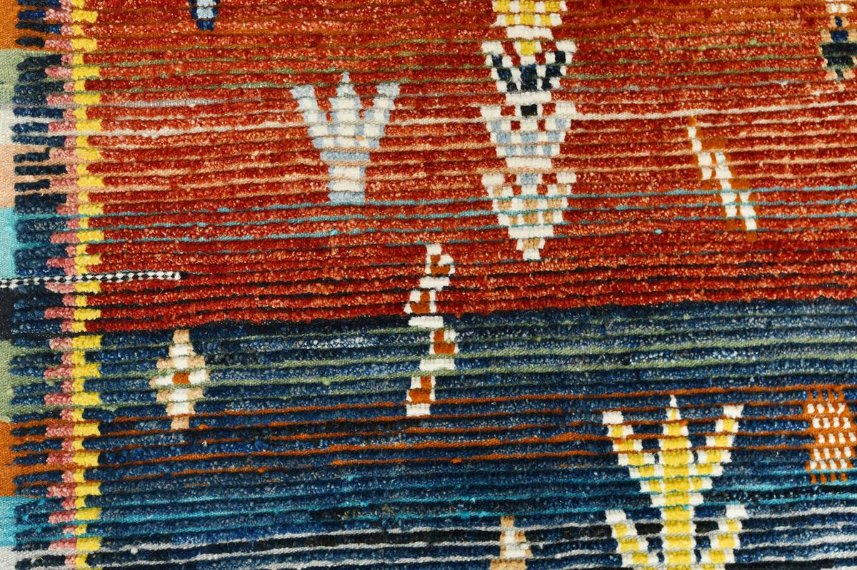 Close-up of a woven rug showing a terracotta red upper section and a dark blue lower section with geometric and floral motifs in cream, yellow, and orange. Atlantica Organics curates antique and handmade rugs, Persian, Afghan, Tibetan, Pakistani, Turkish Oushak, Kilim, Khotan, tribal and contemporary textiles for luxury interiors, homes and real estate. Our collections connect timeless craftsmanship with interior design, furniture, fine art, architecture and modern living. Explore curated rugs, collectible 
