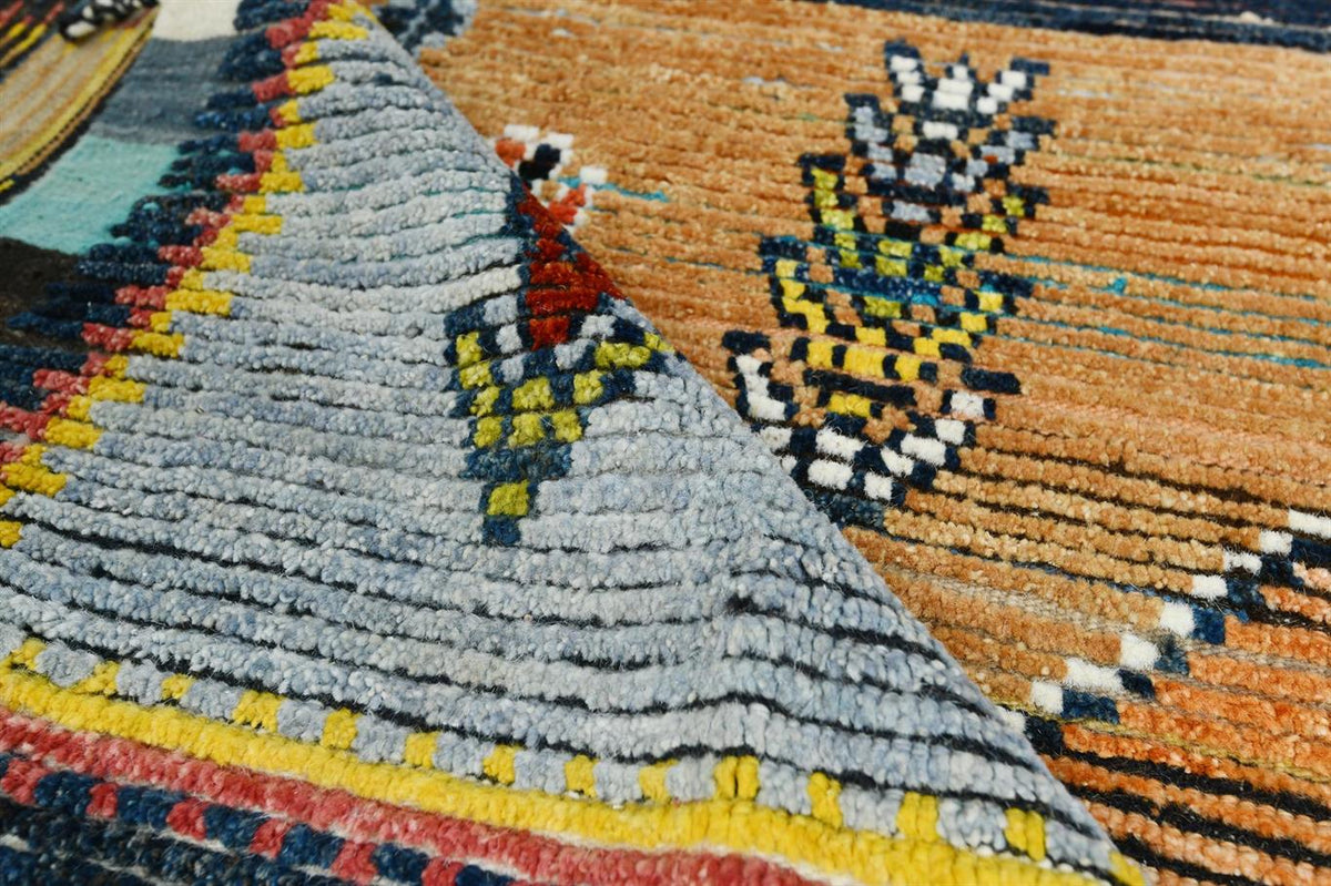 A close-up of a textured, handwoven rug showing two distinct geometric patterns. The left section features a light gray triangular field with a red, yellow, and blue geometric motif, bordered by yellow, red, and blue stripes. Atlantica Organics curates antique and handmade rugs, Persian, Afghan, Tibetan, Pakistani, Turkish Oushak, Kilim, Khotan, tribal and contemporary textiles for luxury interiors, homes and real estate. Our collections connect timeless craftsmanship with interior design, furniture, fine a