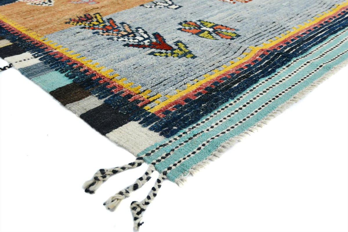 A close-up of a woven rug corner showing a light blue field with multicolored geometric and floral motifs, bordered by black, white, turquoise, yellow, and red stripes, with fringed tassels on a white background. Atlantica Organics curates antique and handmade rugs, Persian, Afghan, Tibetan, Pakistani, Turkish Oushak, Kilim, Khotan, tribal and contemporary textiles for luxury interiors, homes and real estate. Our collections connect timeless craftsmanship with interior design, furniture, fine art, architect