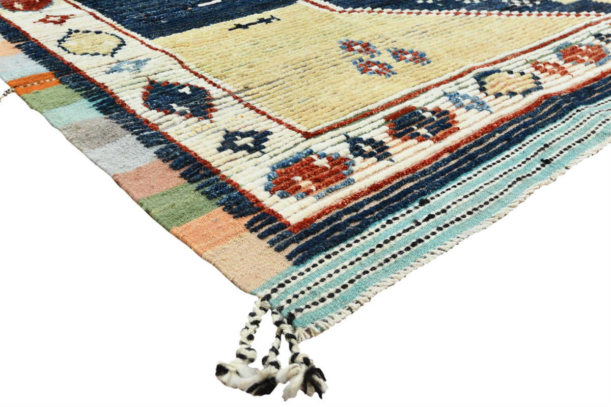 A close-up of a woven Moroccan-style rug corner, showing cream, navy, orange, green, and light blue patterns with braided fringe tassels on a white background. Atlantica Organics curates antique and handmade rugs, Persian, Afghan, Tibetan, Pakistani, Turkish Oushak, Kilim, Khotan, tribal and contemporary textiles for luxury interiors, homes and real estate. Our collections connect timeless craftsmanship with interior design, furniture, fine art, architecture and modern living. Explore curated rugs, collecti